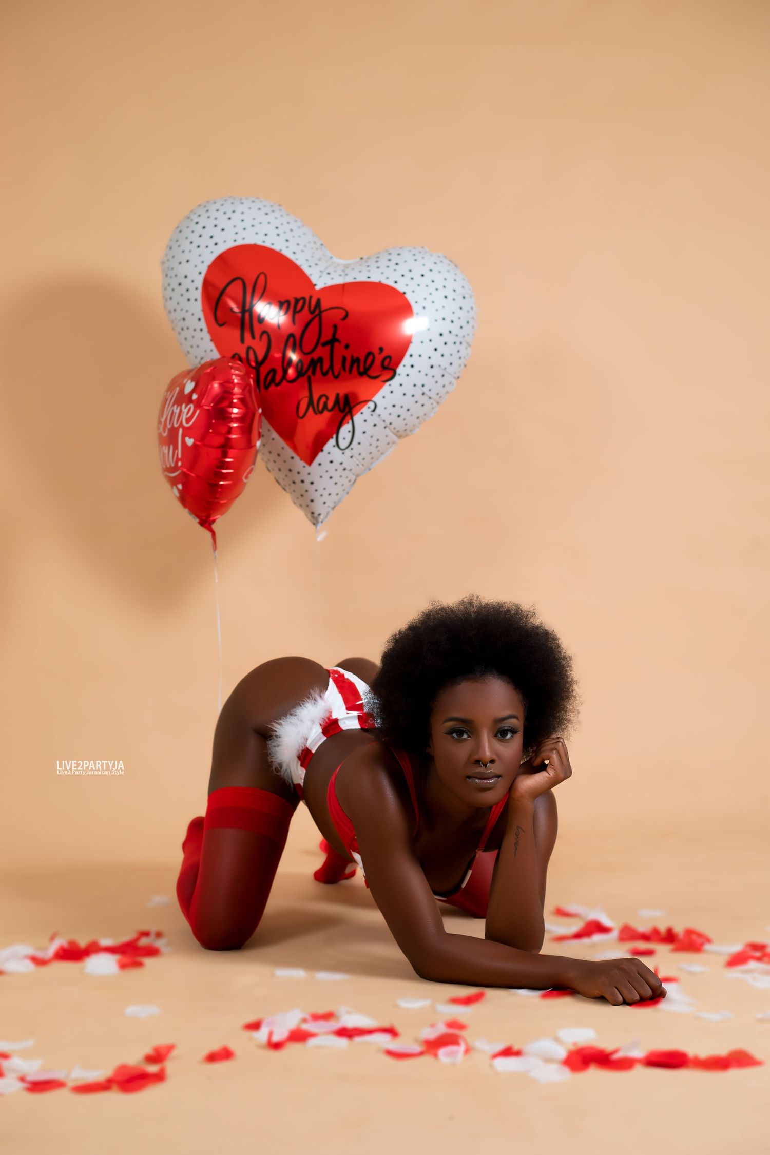 Woman in red lingerie poses with Happy Valentines Day heart balloons on beige background with rose petals.