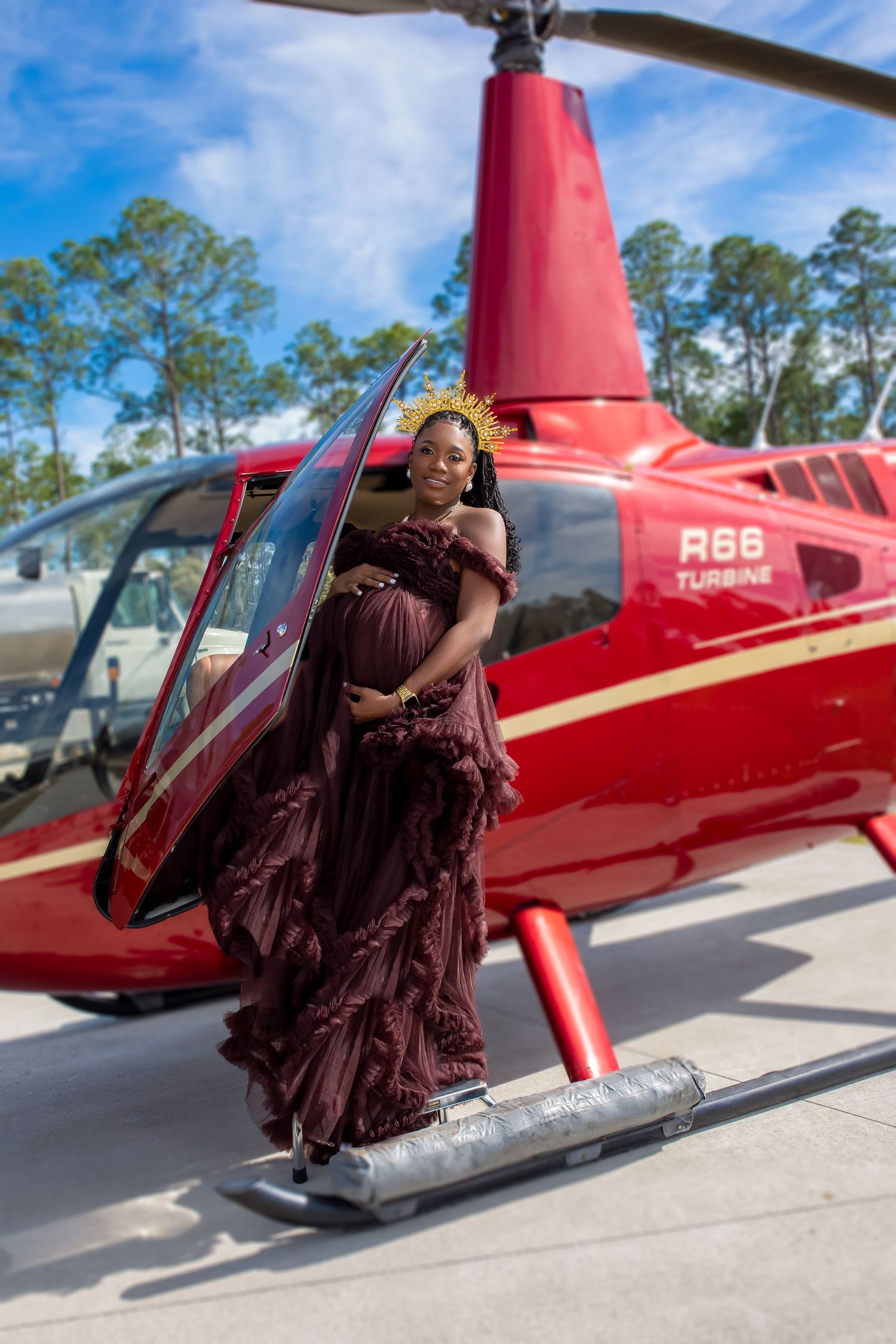 Smiling pregnant woman in burgundy ruffle dress with gold crown posing beside red R66 helicopter.