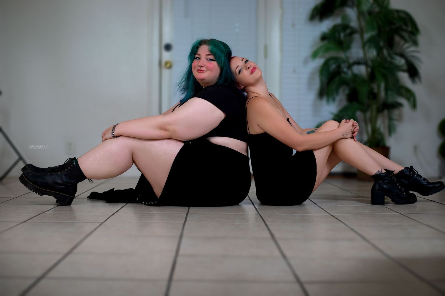 Two women in black outfits sitting back to back on tile floor.