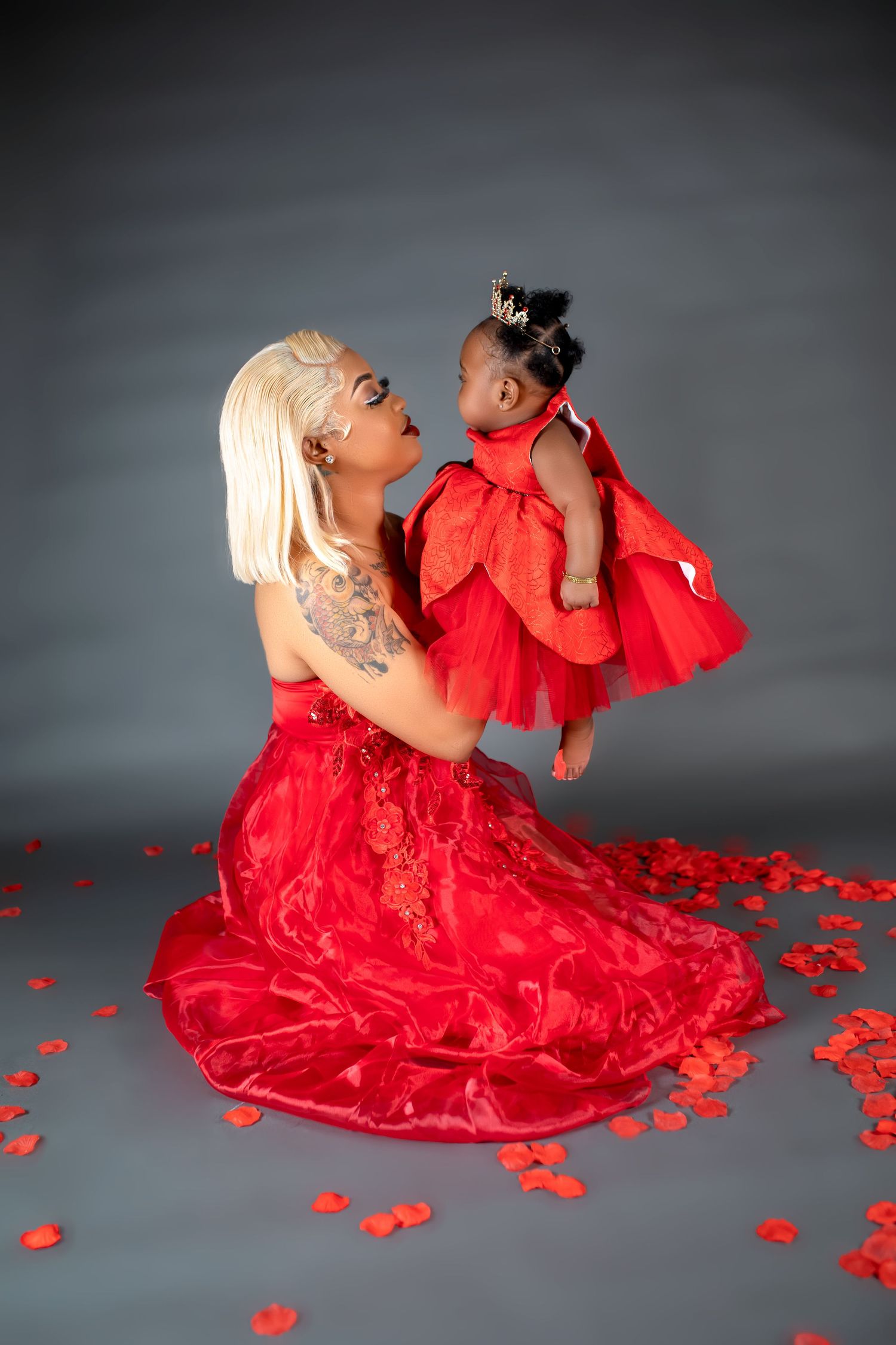 Woman in red gown holds baby girl in matching red tulle dress surrounded by rose petals.