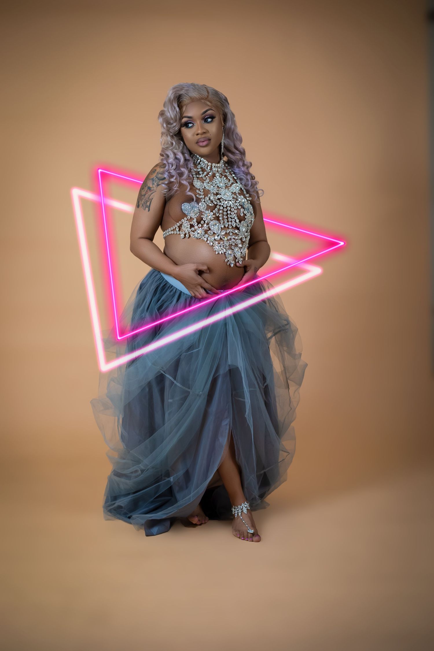 Pregnant woman in lace crop top and blue tulle skirt poses with pink neon triangle on beige background.