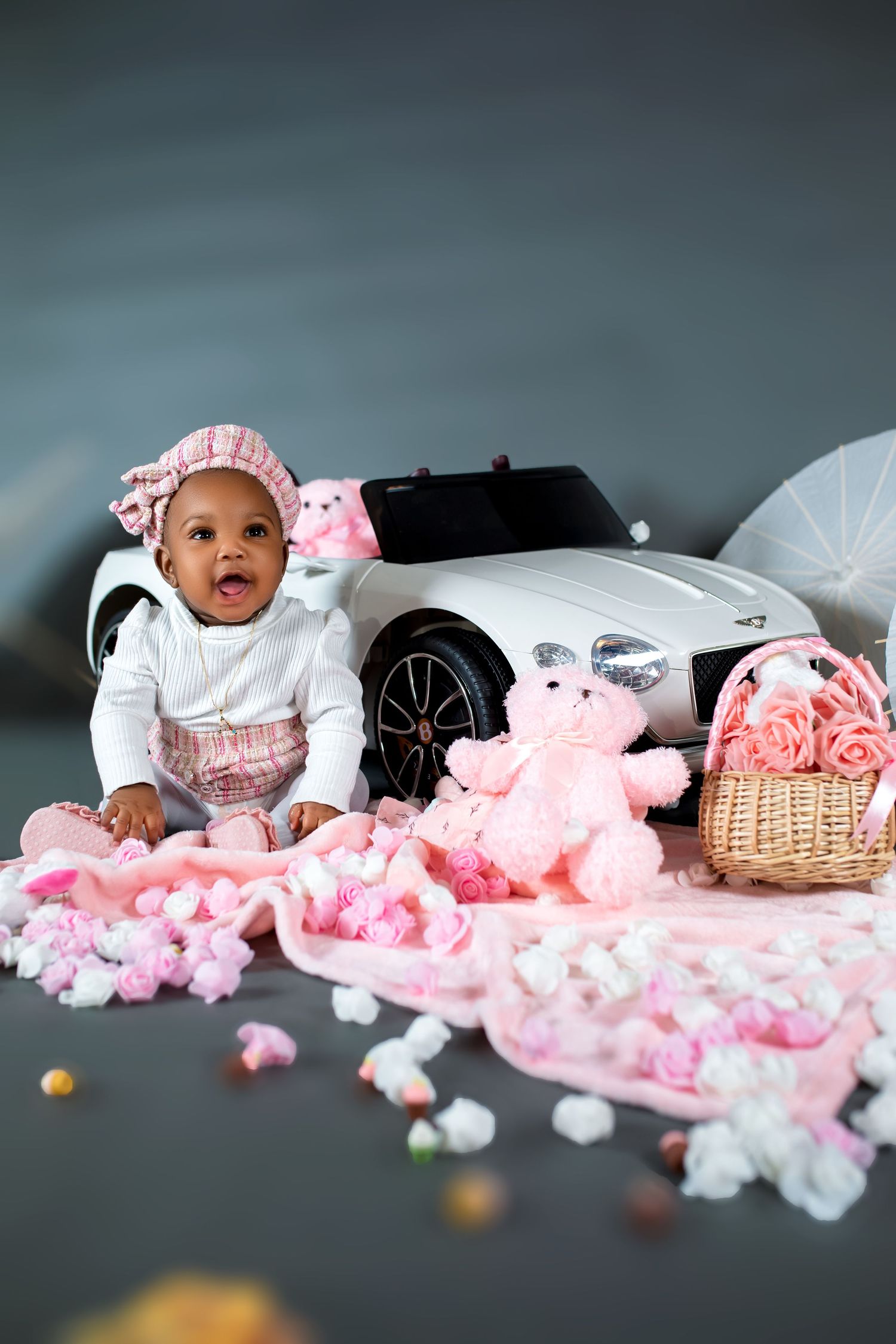 Adorable baby girl in pink outfit sits beside toy car with teddy bears and pink flowers.