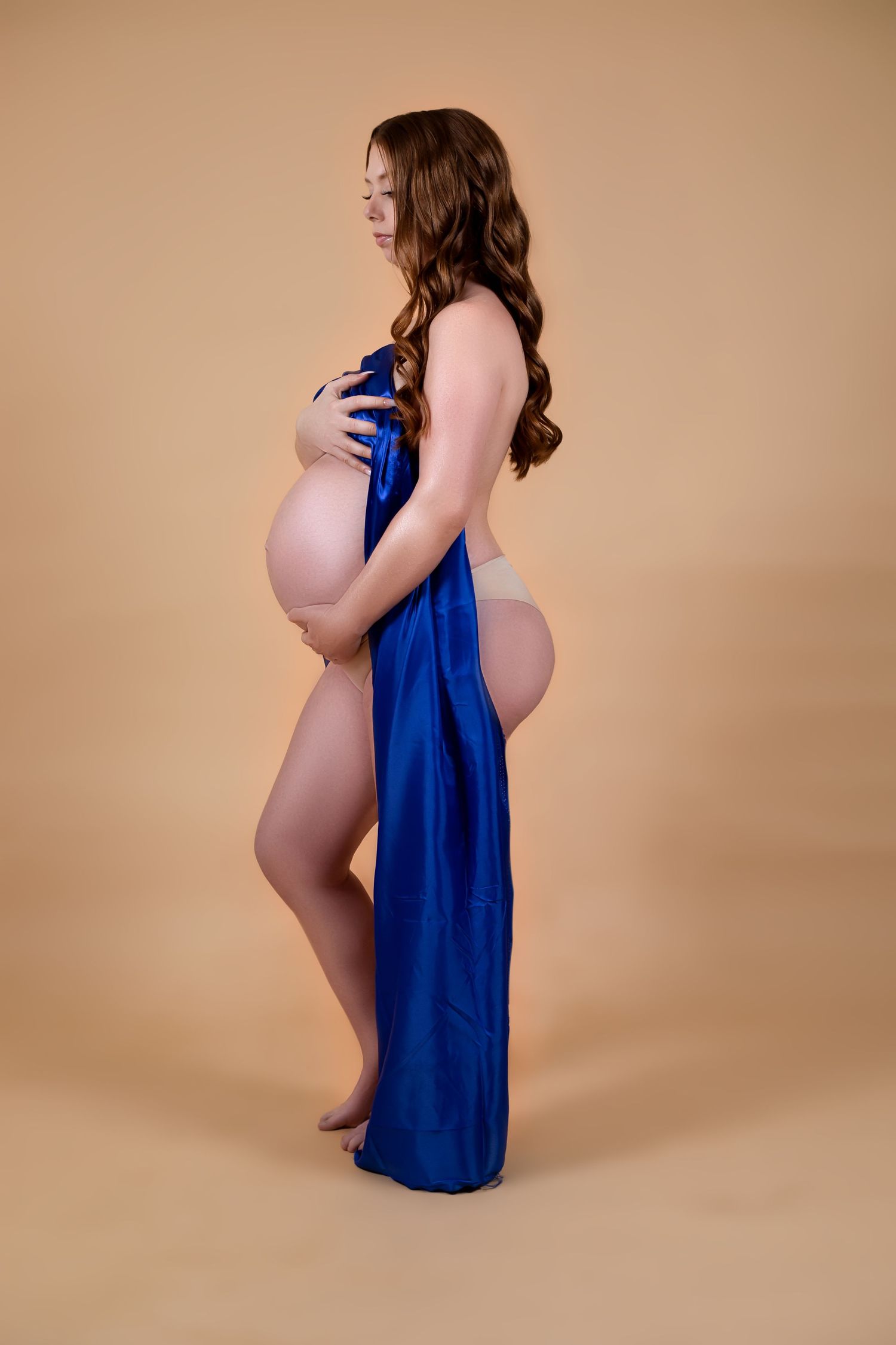 Pregnant woman in blue satin wrap posing sideways on beige studio background showcasing baby bump.