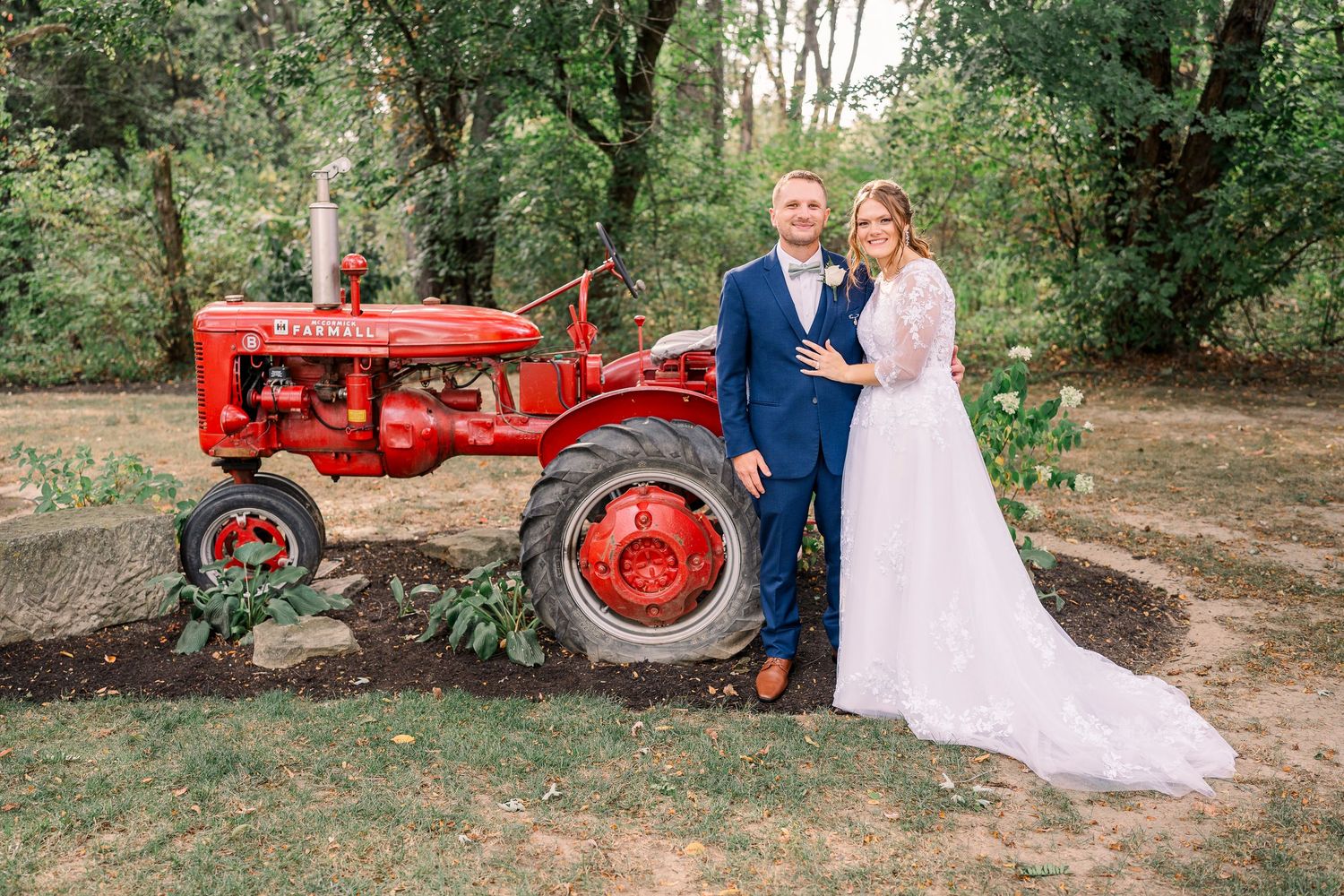 Bride and groom posing beside a vintage red Farmall tractor in a lush garden setting at Gervasi Vineyard wedding venue.
