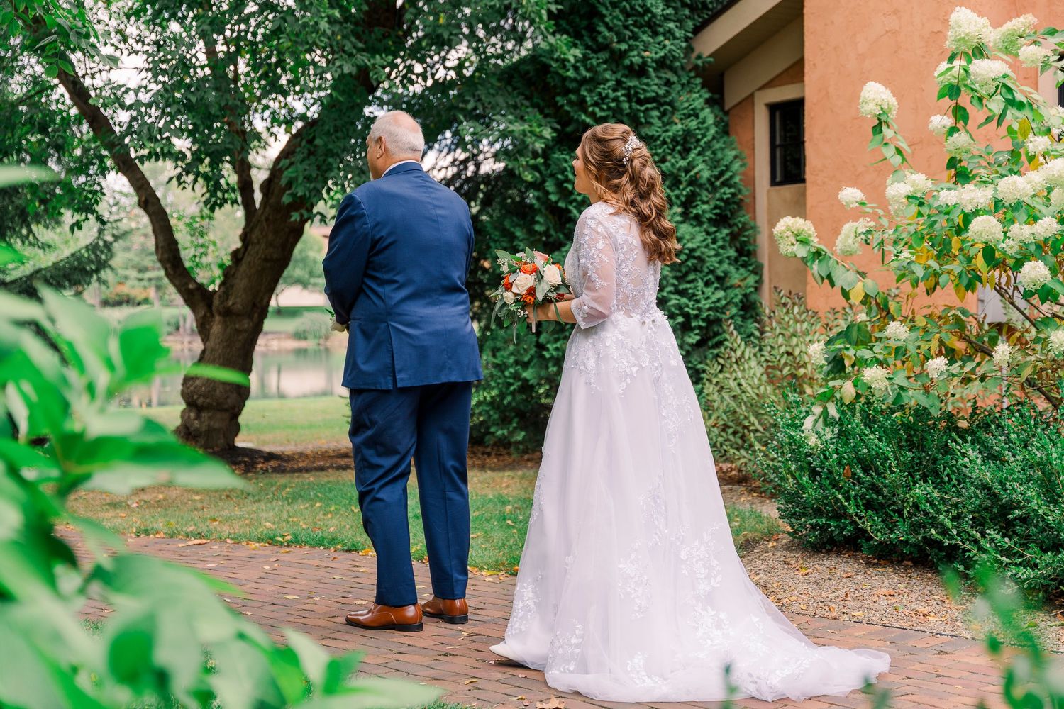 Bride in lace gown and groom in blue suit walking on brick garden path.