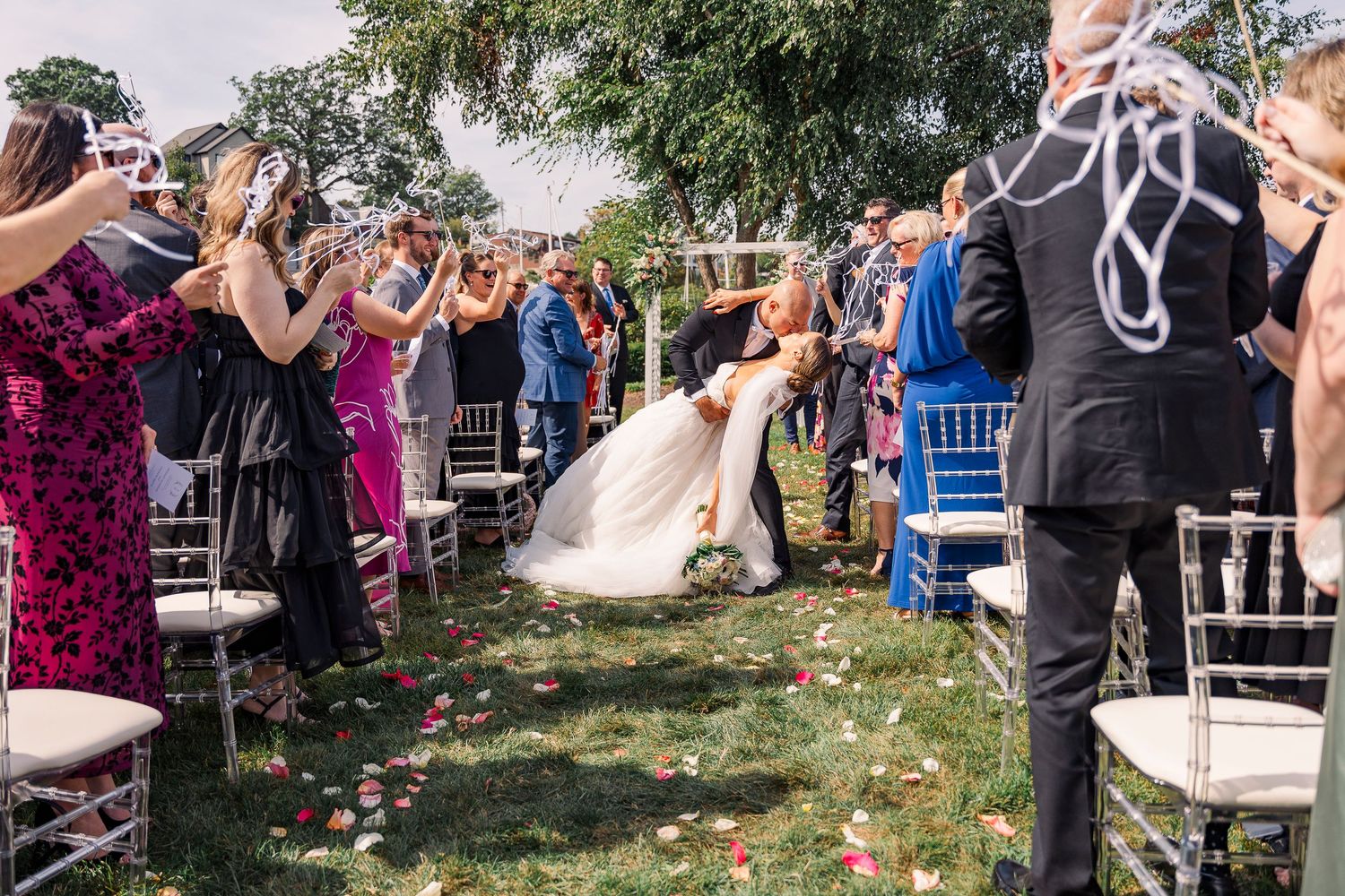 Bride and groom share a kiss at outdoor wedding ceremony while guests cheer along rose petal lined aisle.