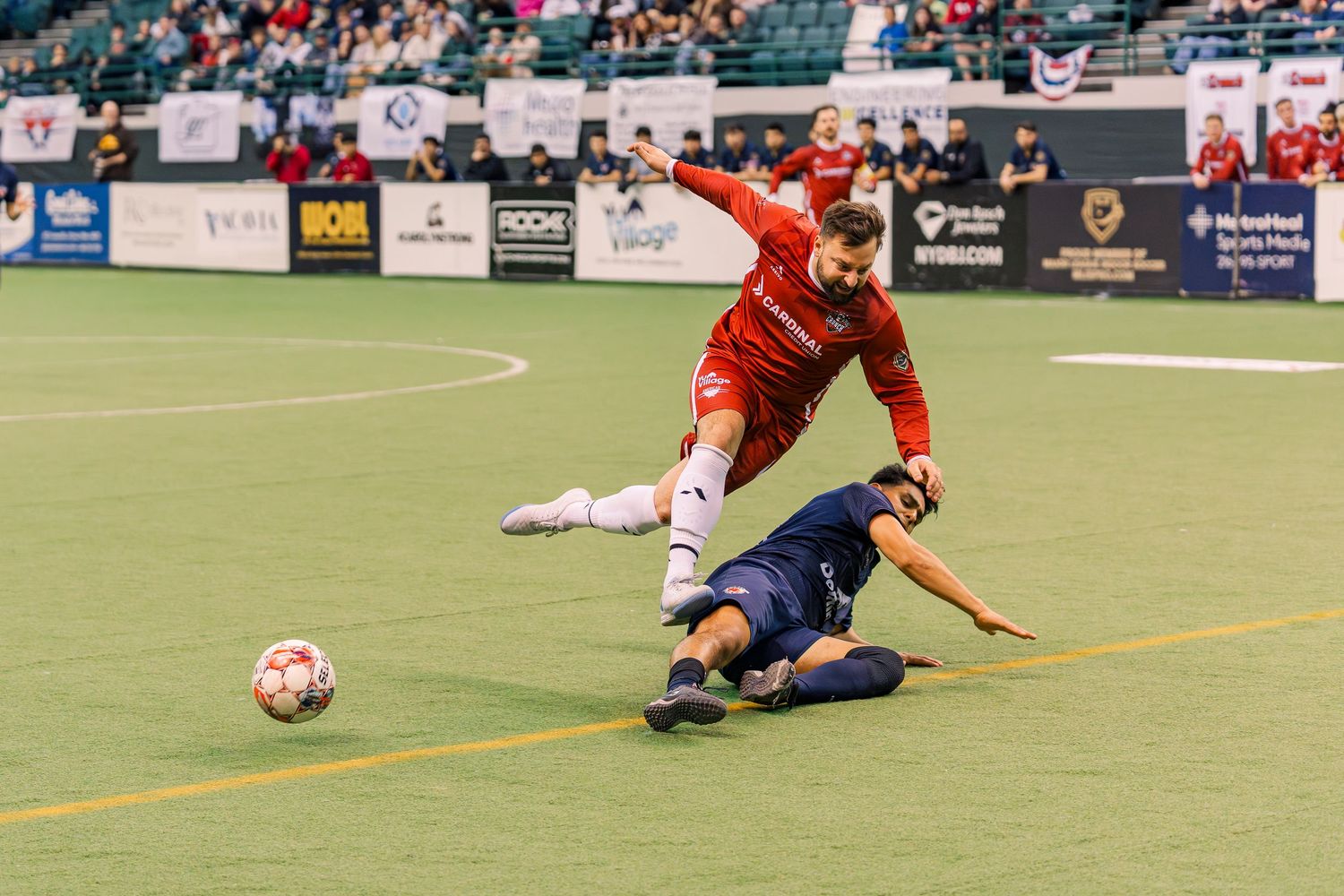 Two soccer players compete in an indoor arena, one in red sliding tackling the other in a dark uniform near the ball. Cleveland Crunch game.