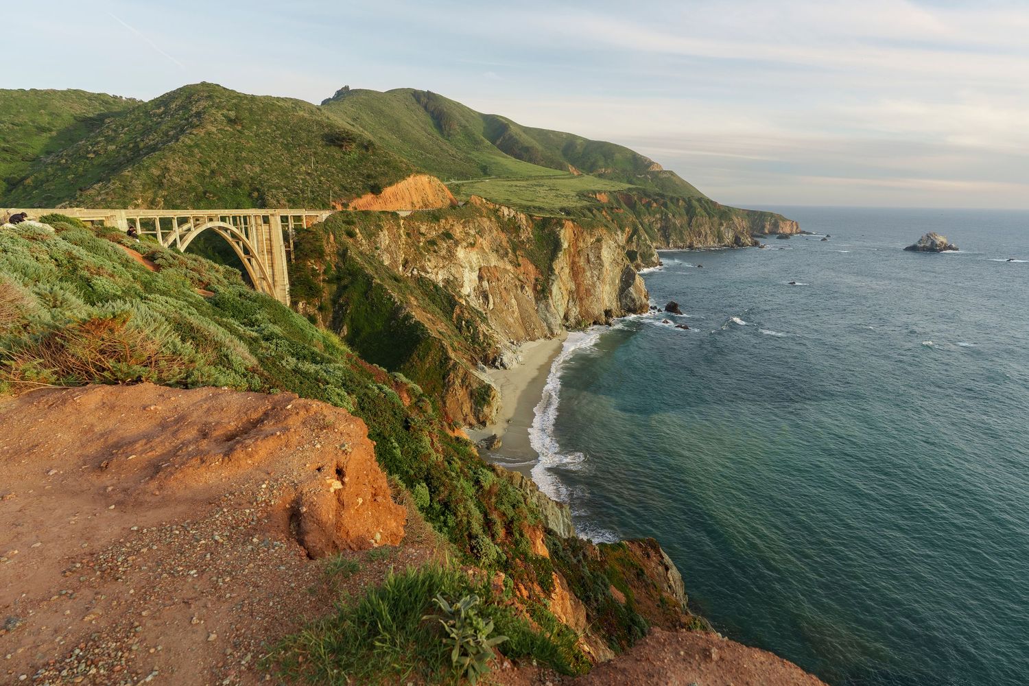 Bixby Creek Bridge arches over rugged Big Sur coastline with green hills, dramatic cliffs, and Pacific Ocean views in California.