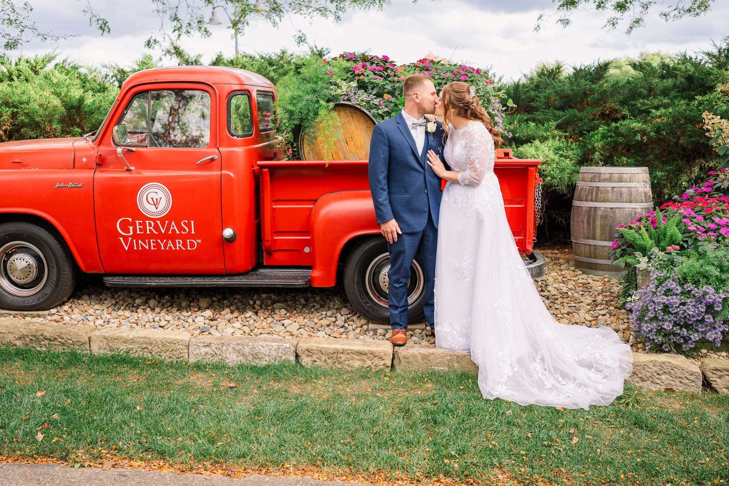 Bride and groom kissing beside a vintage red Gervasi Vineyard truck surrounded by wine barrels.