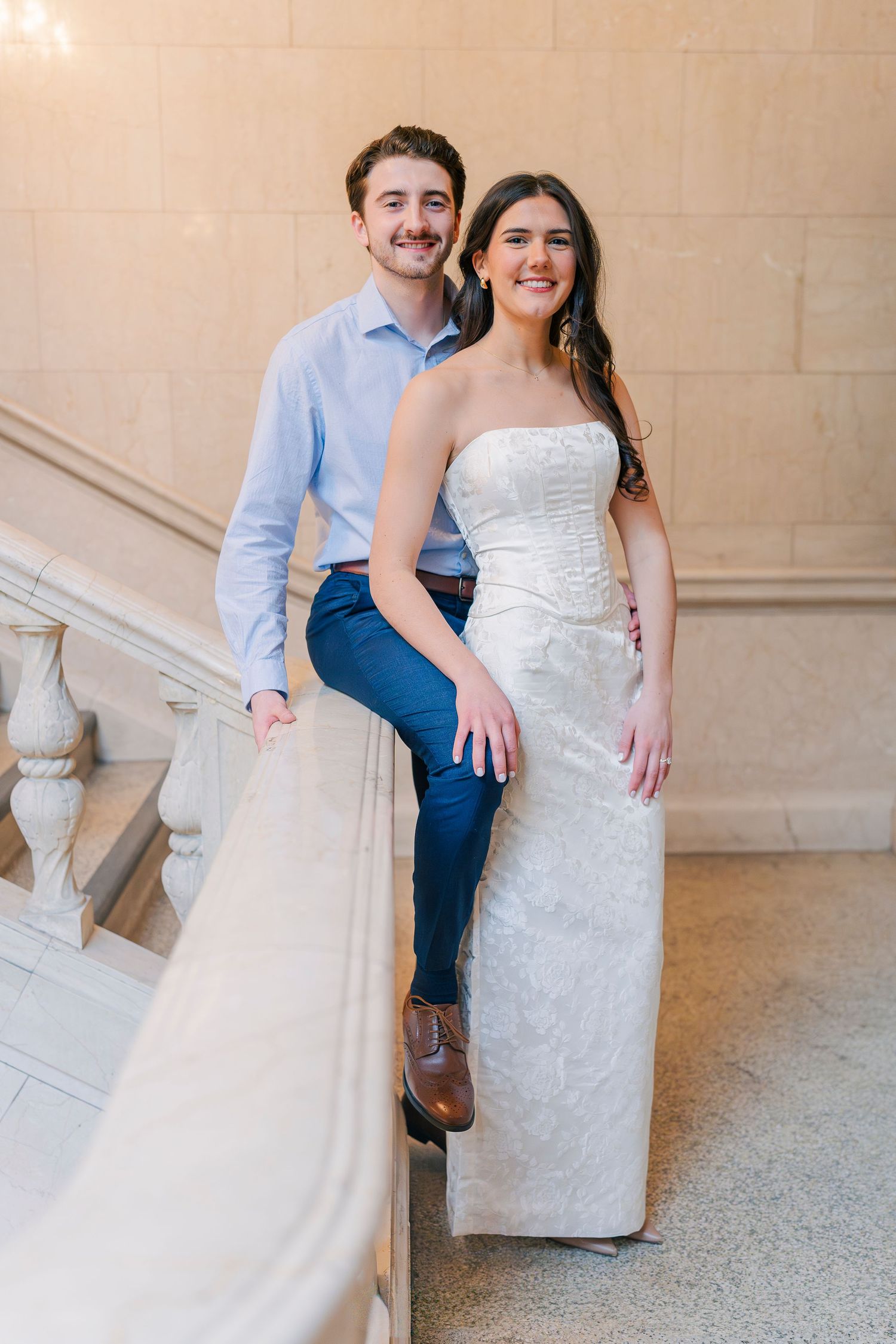 A happy couple poses on marble stairs, the woman in a white strapless gown and the man in a light blue shirt at the Cleveland Public Library