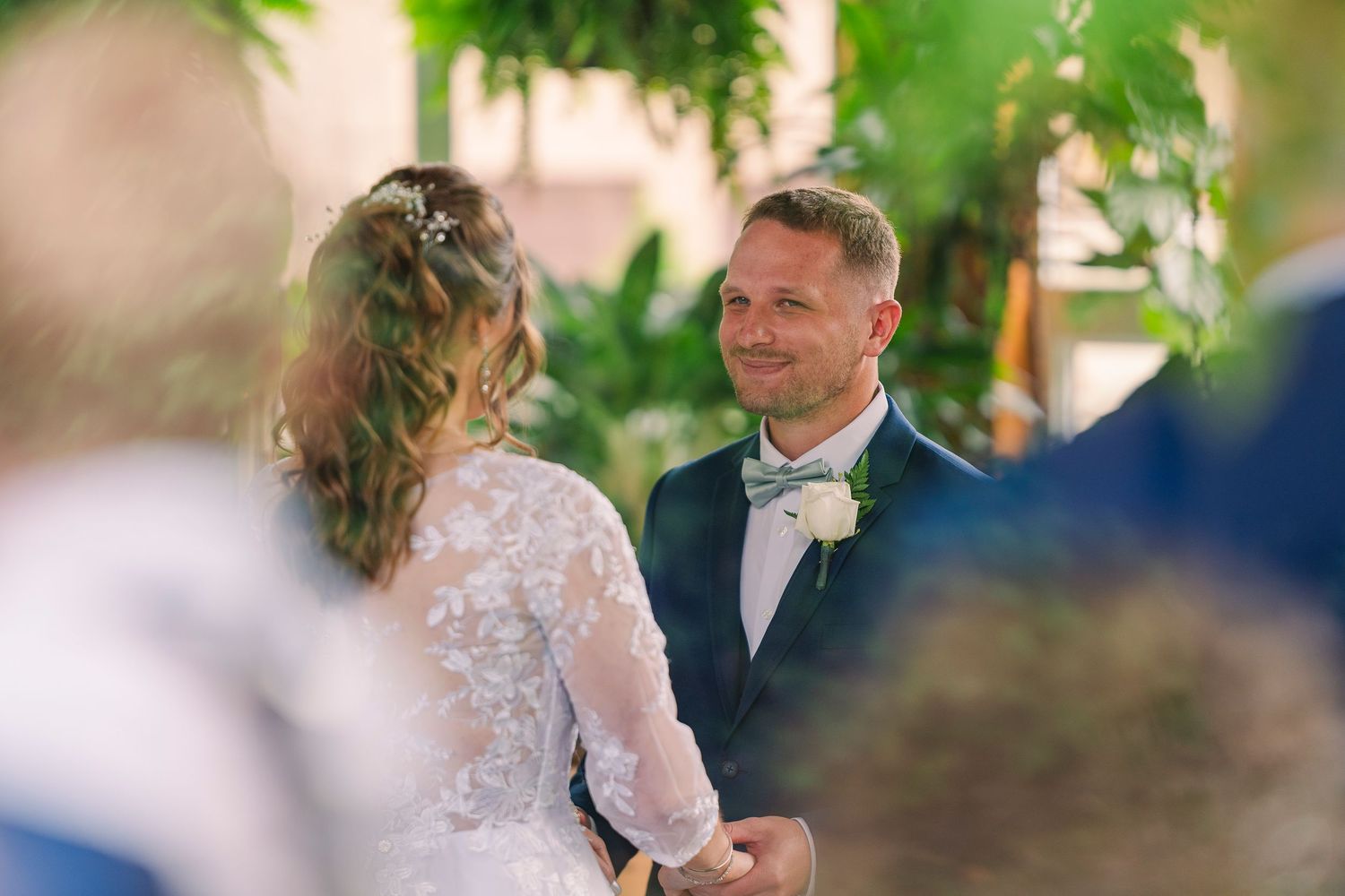Groom in navy suit smiling at bride during romantic outdoor wedding ceremony.