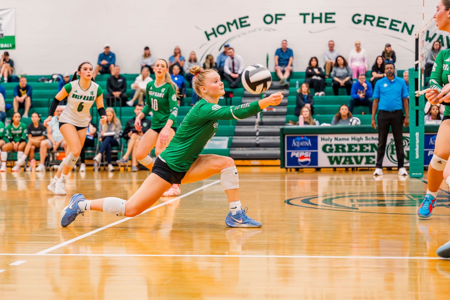 Holy Name volleyball player diving to dig the ball during a high school match in a green and white gymnasium.