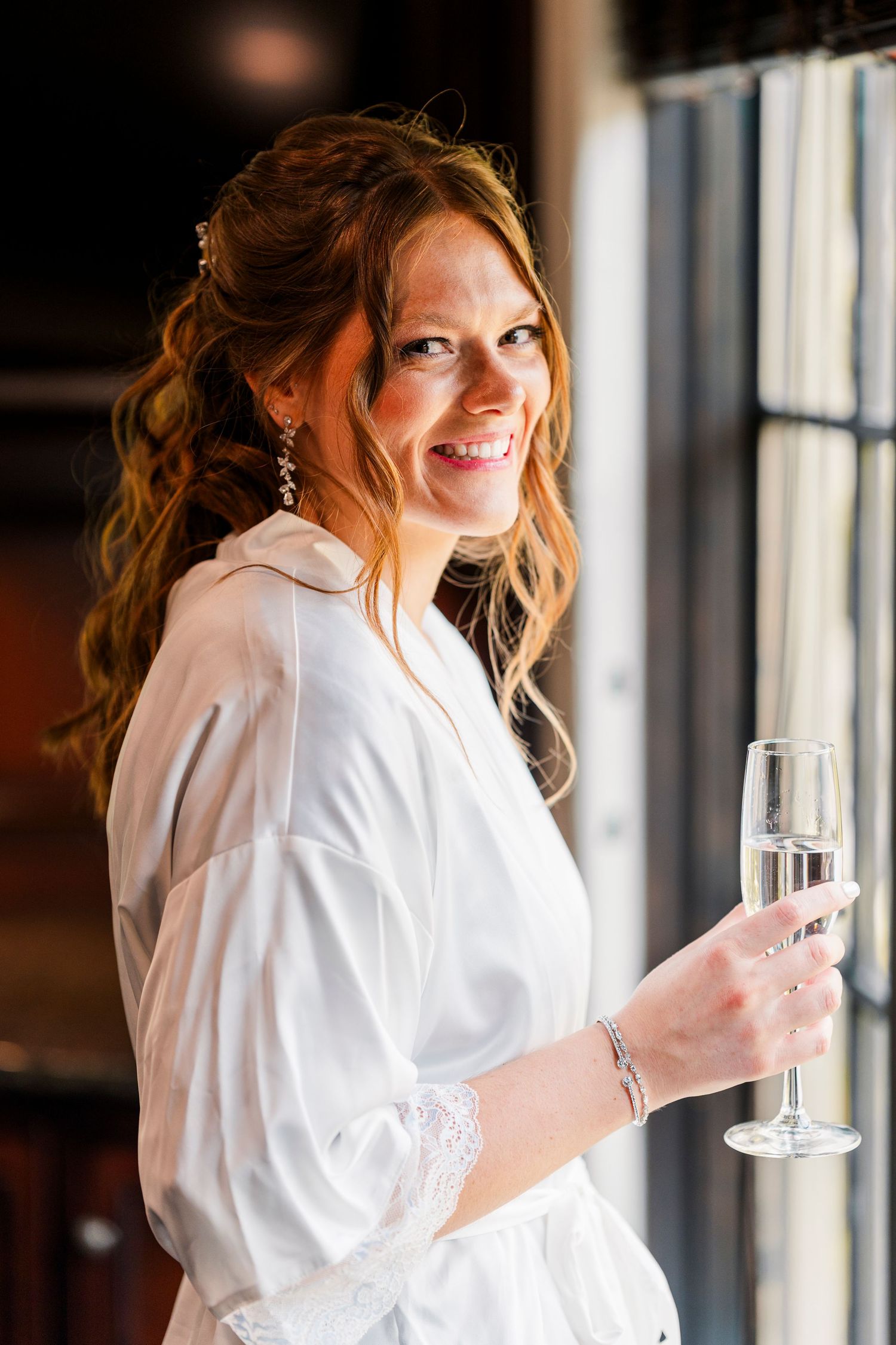Smiling bride in white robe holding champagne flute by window on wedding day.
