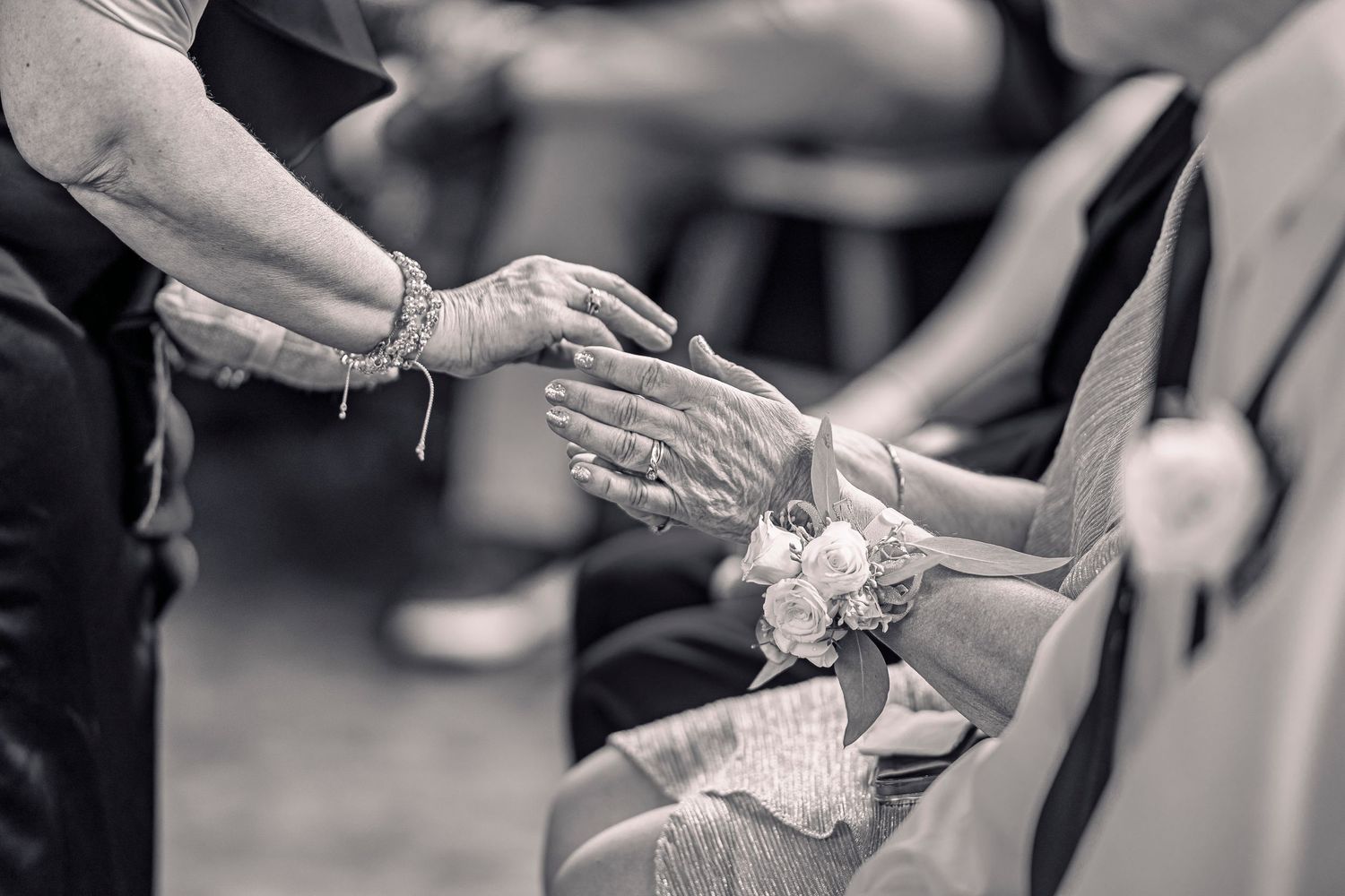 Black and white image of elderly wedding guests holding hands with floral corsage during warming of the rings ceremony