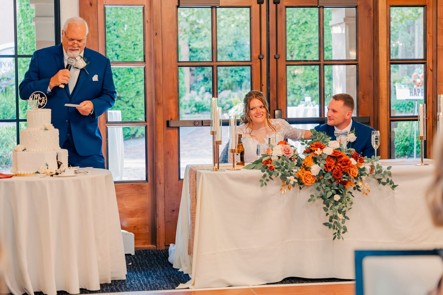 Wedding guest delivers heartfelt speech to smiling bride and groom seated at decorated reception table.