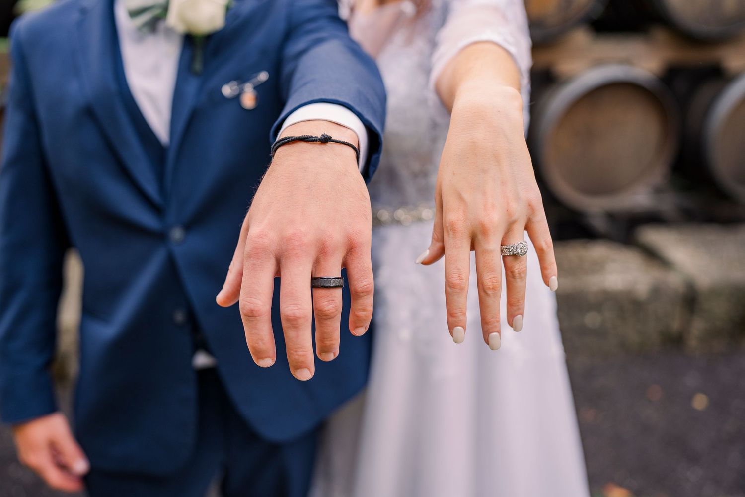Newlyweds displaying their wedding rings with wine barrels in the background at a vineyard.