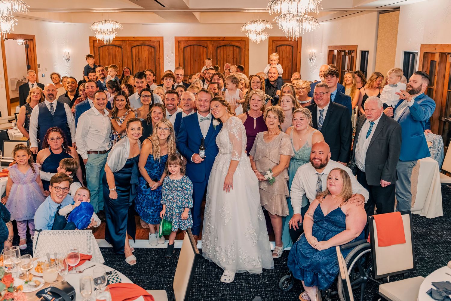 Large wedding party group photo with bride and groom surrounded by guests in decorated reception hall.