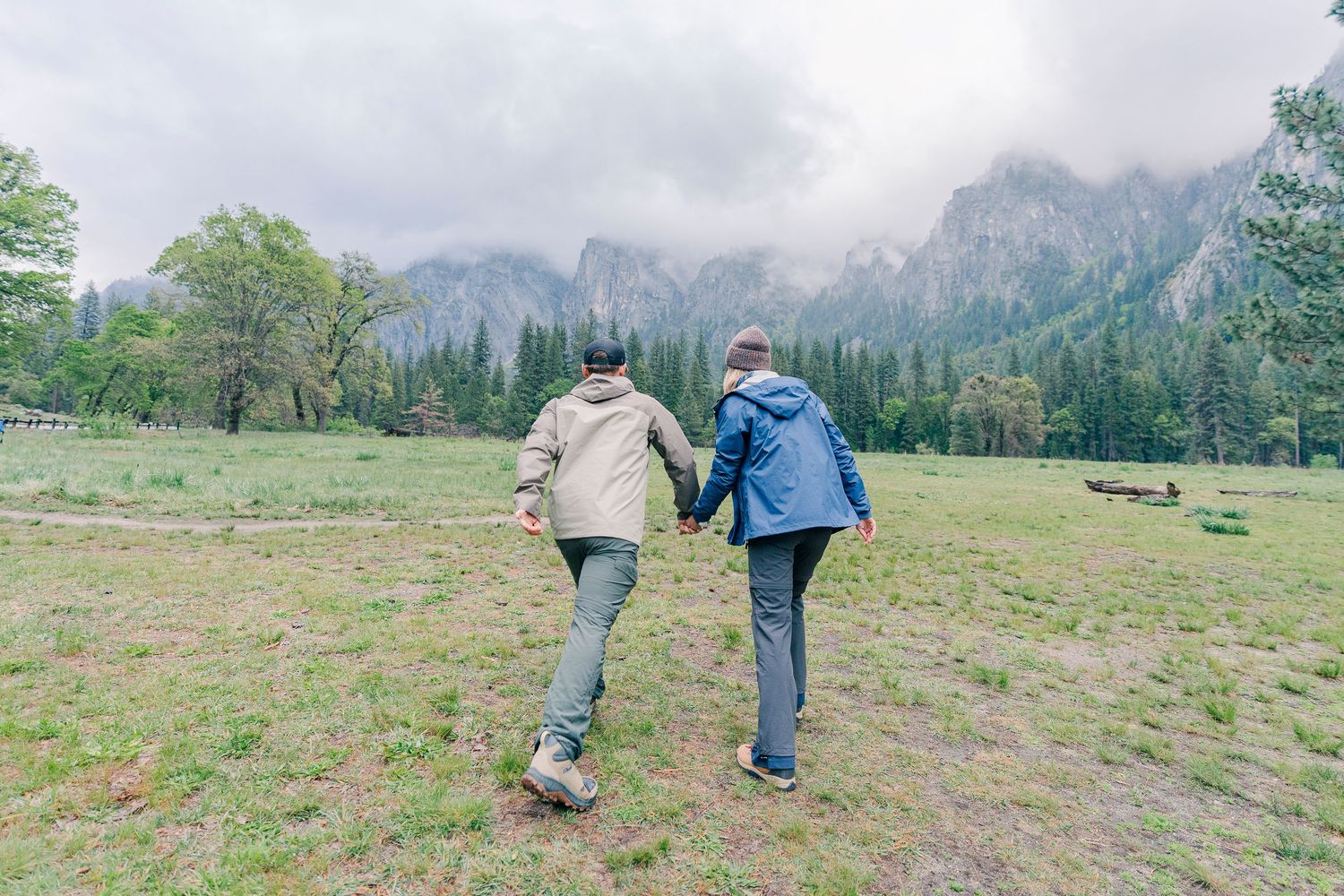 Couple holding hands walking through a lush green meadow with pine trees and misty mountains in the background at Yosemite National Park