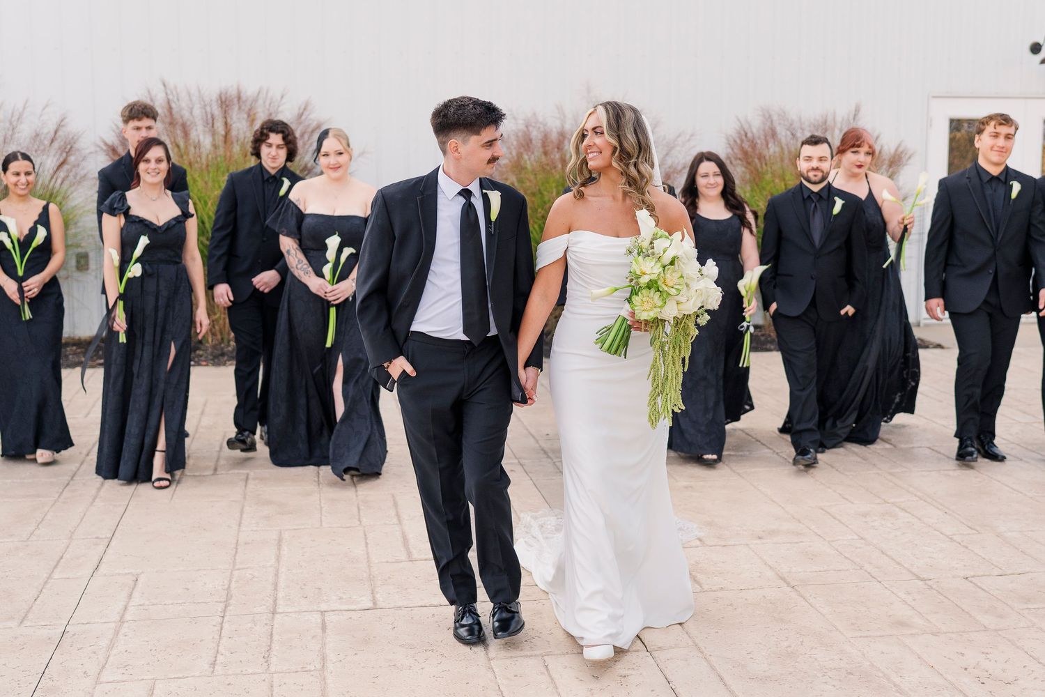 Bride and groom walk hand-in-hand with wedding party in black attire, bride holding white bouquet outdoors.