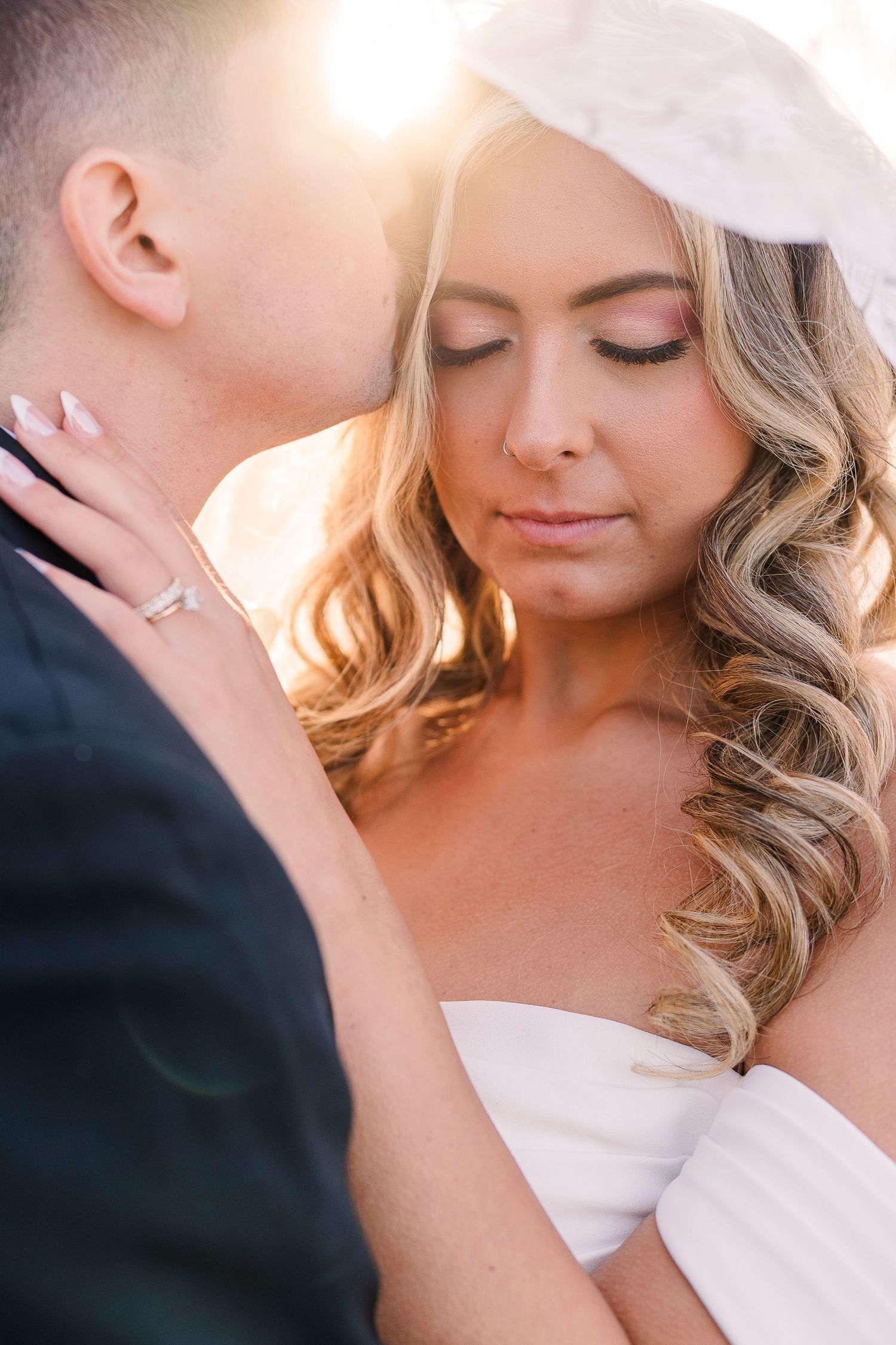 Bride and groom share a tender moment in golden backlight, bride wearing white dress and veil with elegant curled hair.