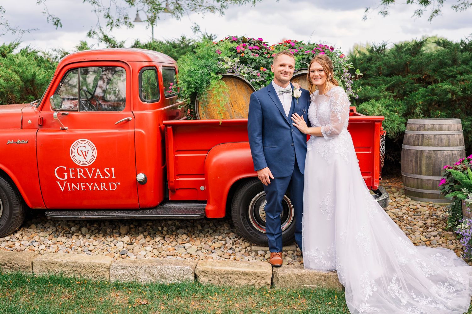 Bride and groom posing beside a vintage red Gervasi Vineyard truck surrounded by lush greenery.