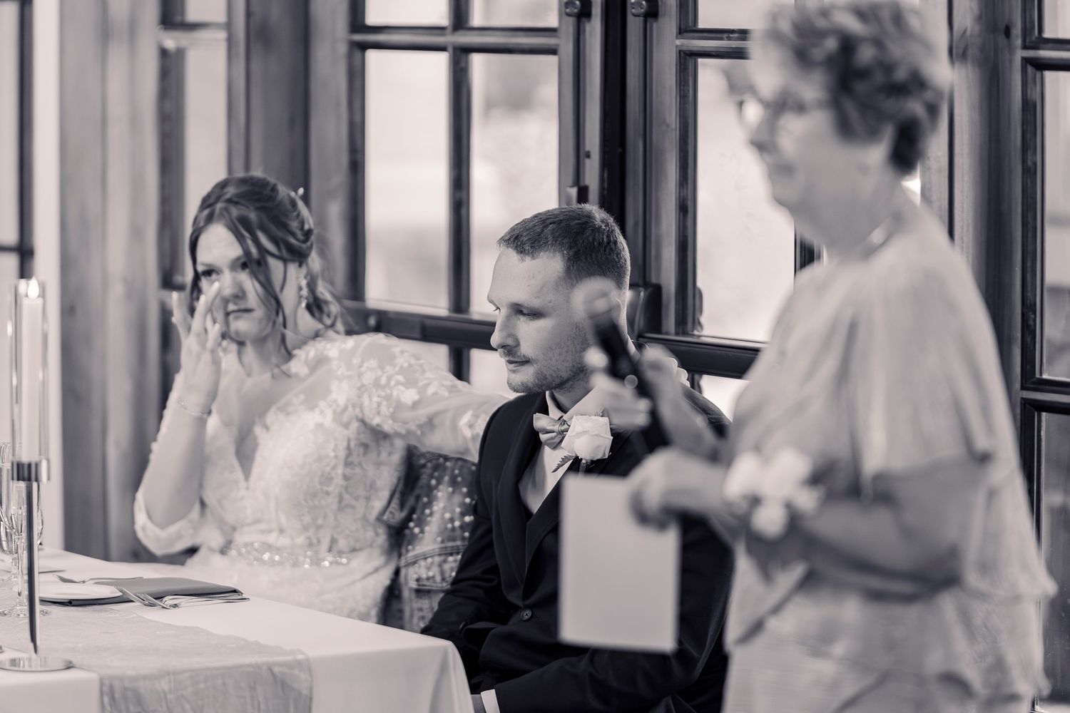 Bride and groom listen emotionally to a wedding speech at their reception table in black and white photo.
