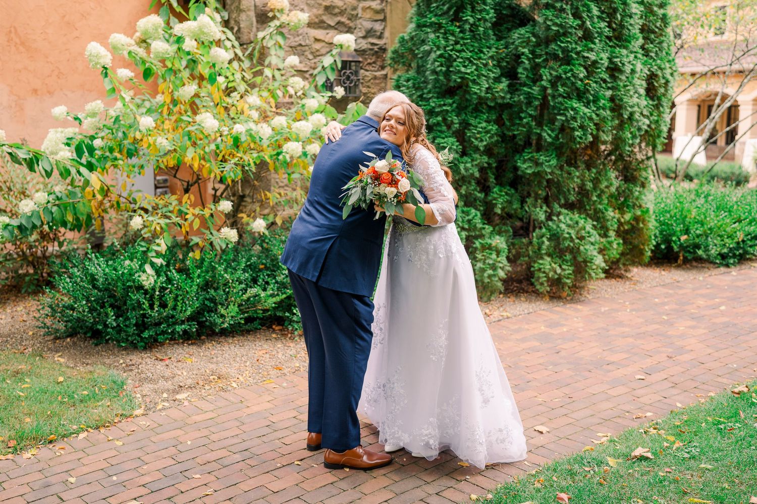 Bride in white lace gown embraces man in navy suit on brick path beside stone building and garden.