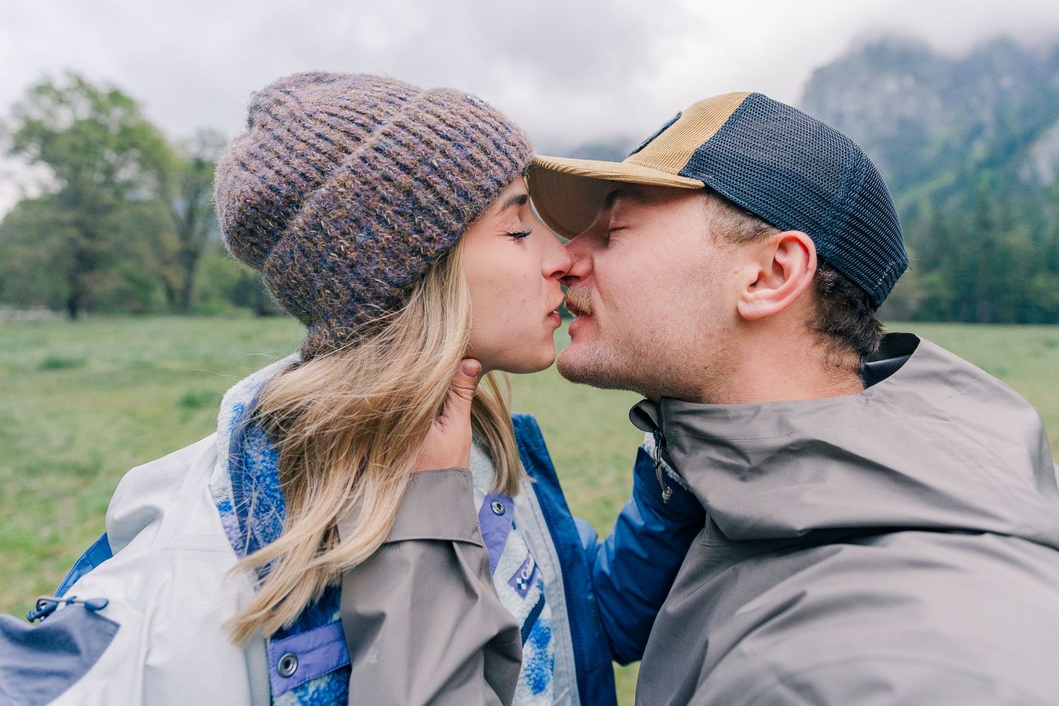Couple sharing a kiss outdoors in a green meadow, wearing winter jackets and hats with mountains in background in Yosemite National Park