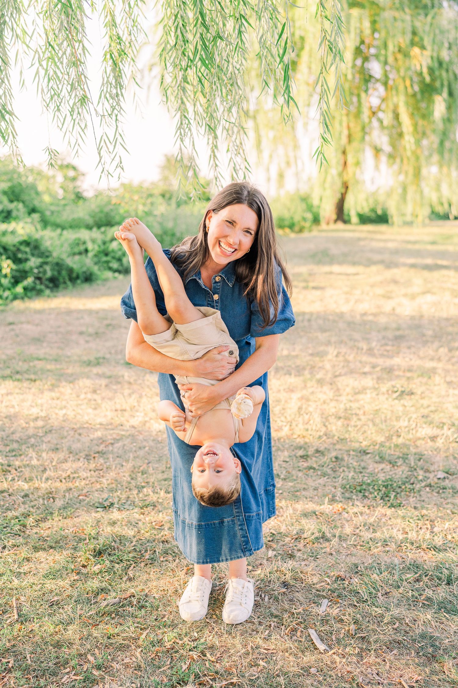 Smiling mother in blue dress playfully holds her toddler upside down under a weeping willow tree at Edgewater Park in Cleveland, Ohio