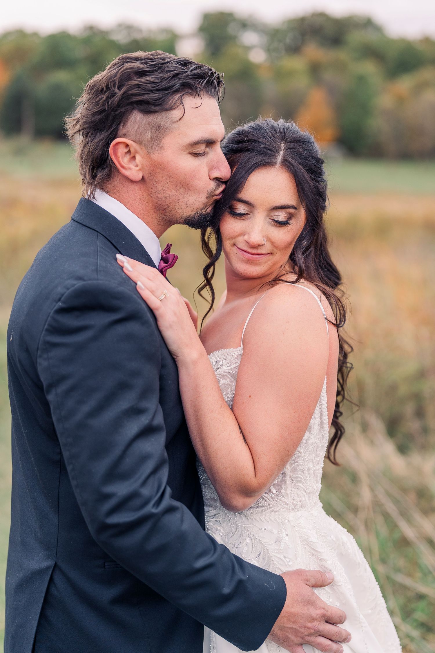 Groom in navy suit kisses bride on forehead in romantic outdoor fall wedding portrait.