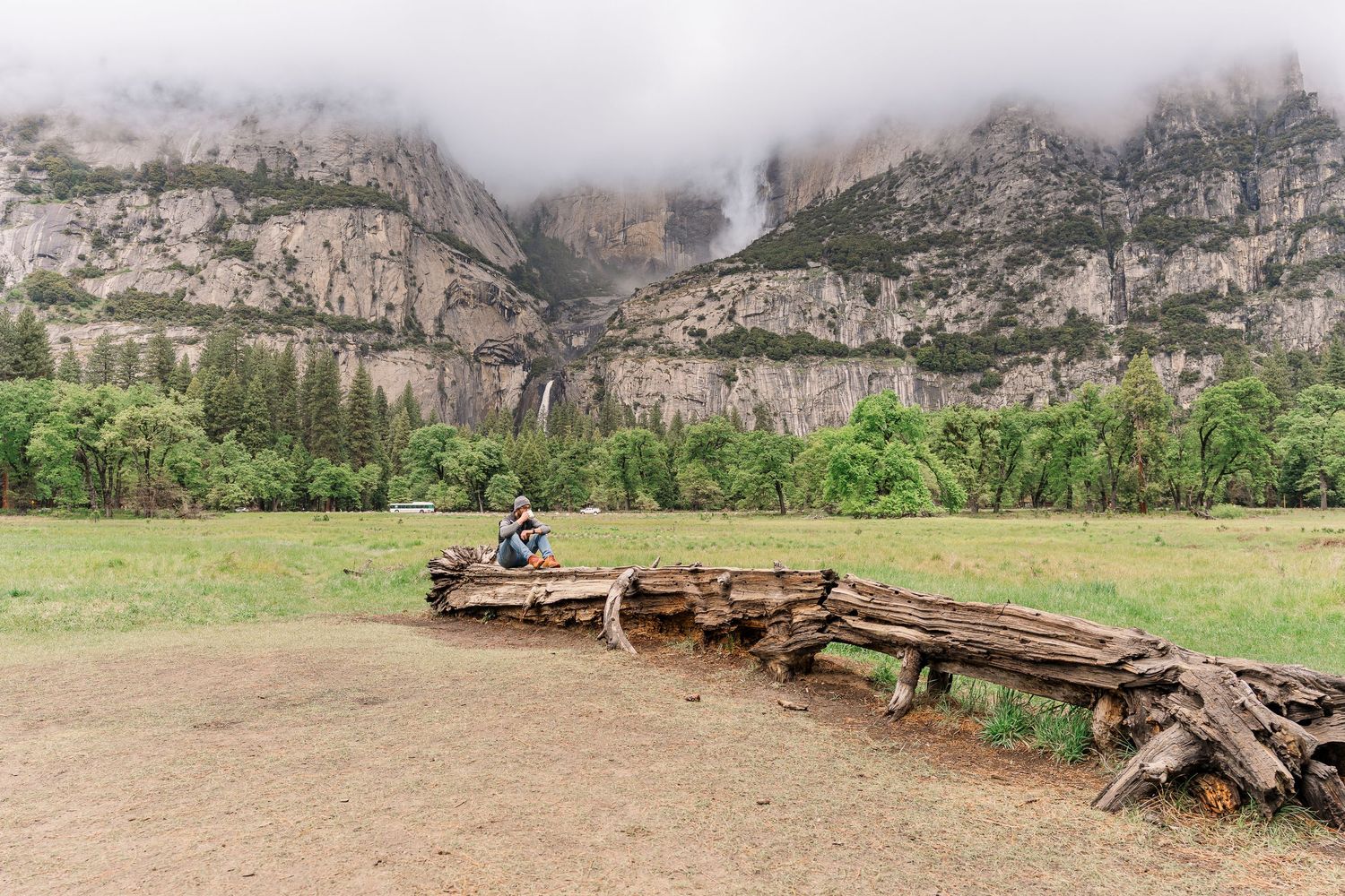 Person sits on fallen logs in Yosemite Valley with waterfall and misty cliffs in background.