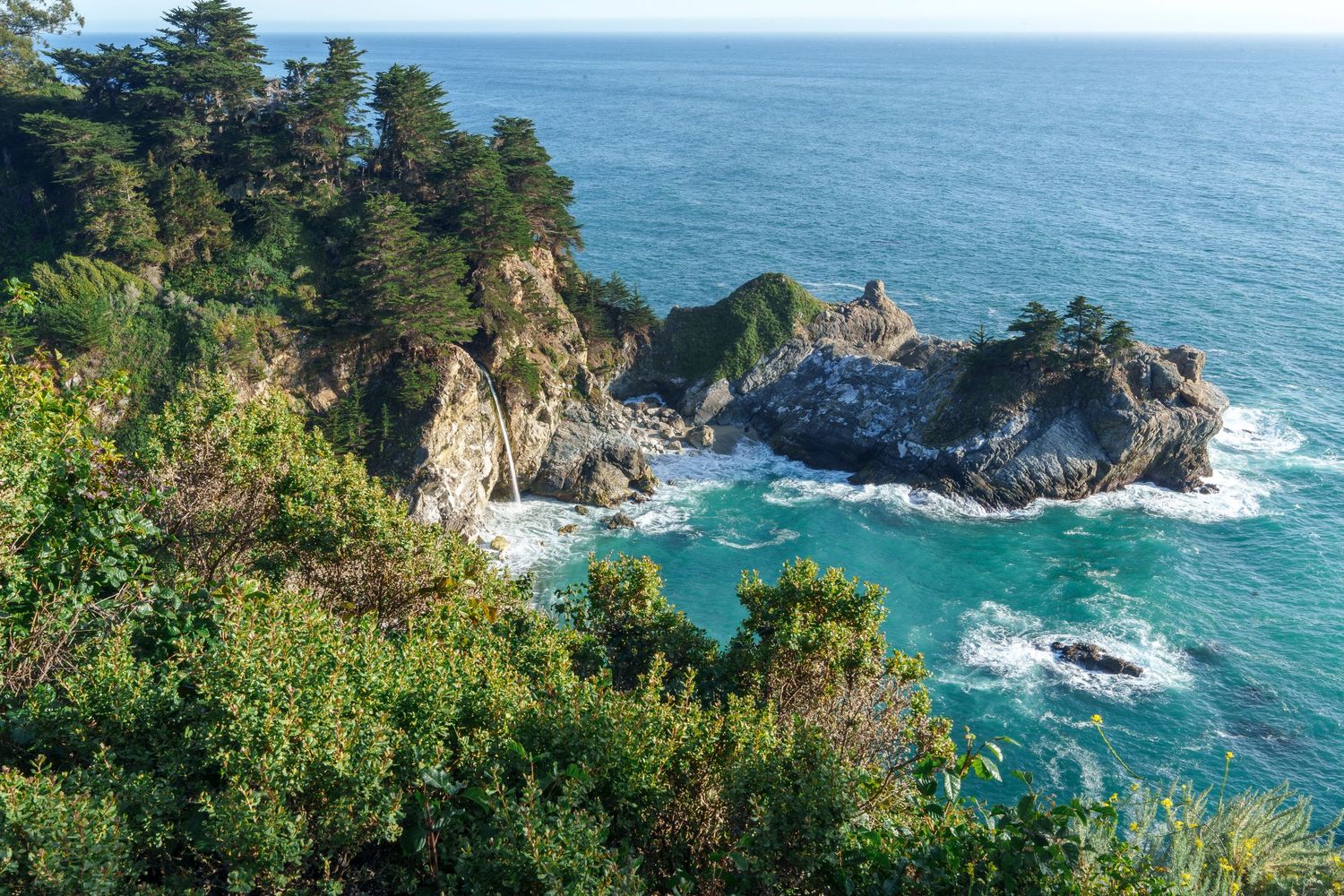 Aerial view of rocky coastal cliffs with lush green trees overlooking turquoise ocean waters at Big Sur, California.