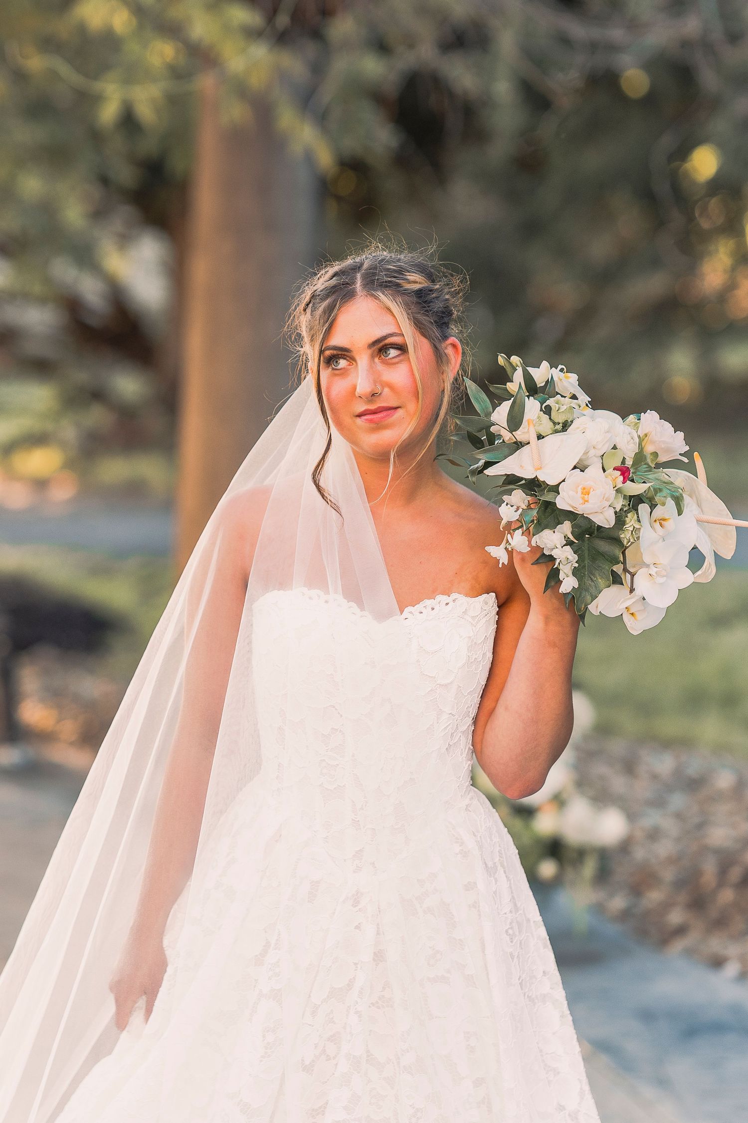 Bride in lace ball gown with veil holding floral bouquet while gazing upward in golden afternoon light.