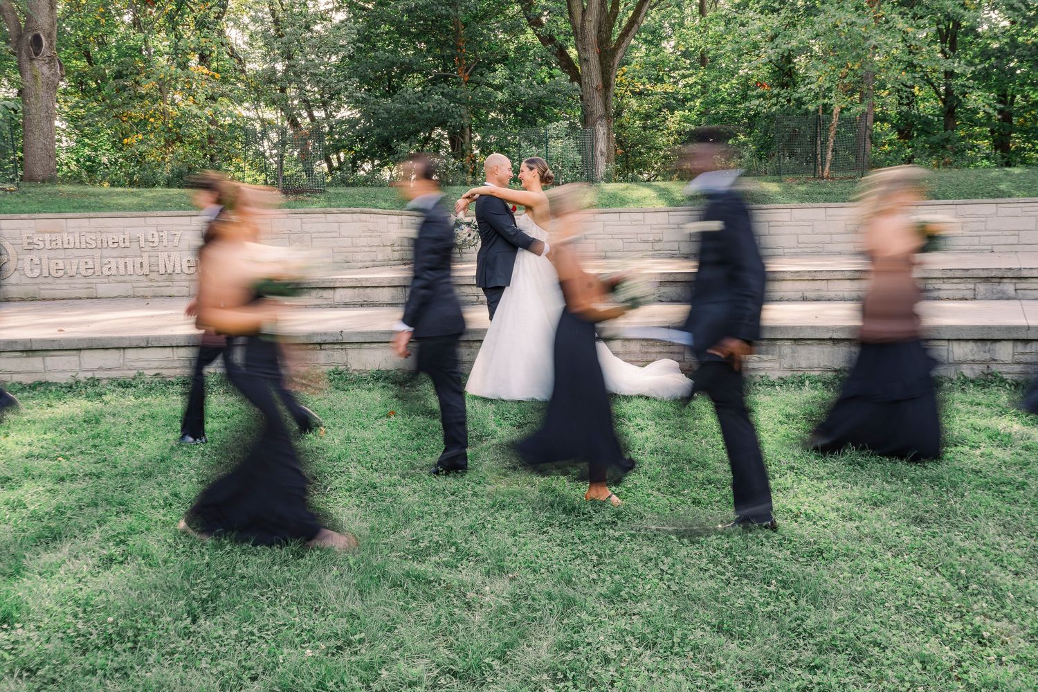 Bride and groom embrace outdoors as blurred wedding party walks past on grass near stone steps surrounded by trees.