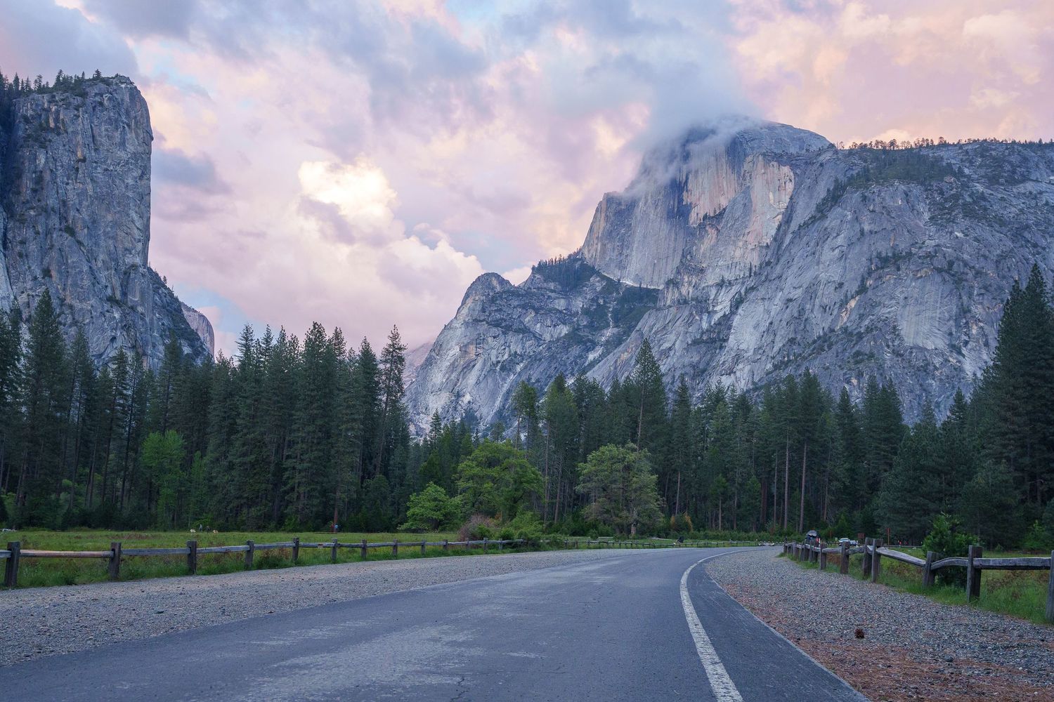 A winding road leads through Yosemite Valley with towering granite cliffs and pine trees under a dramatic sunset sky.