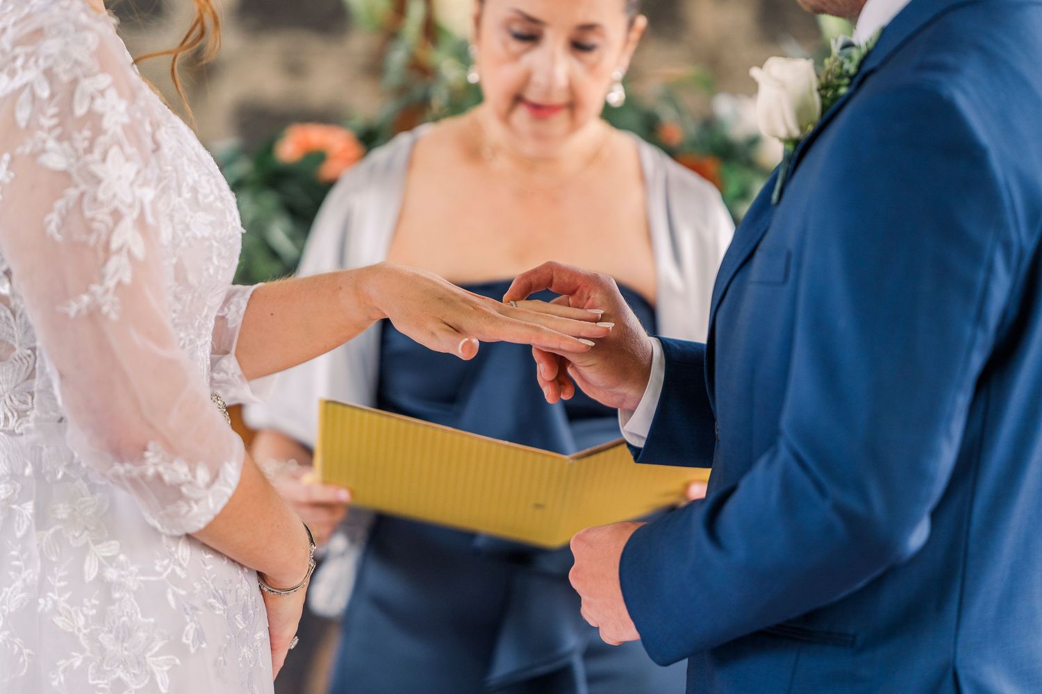 Bride placing wedding ring on grooms finger during ceremony with officiant smiling.