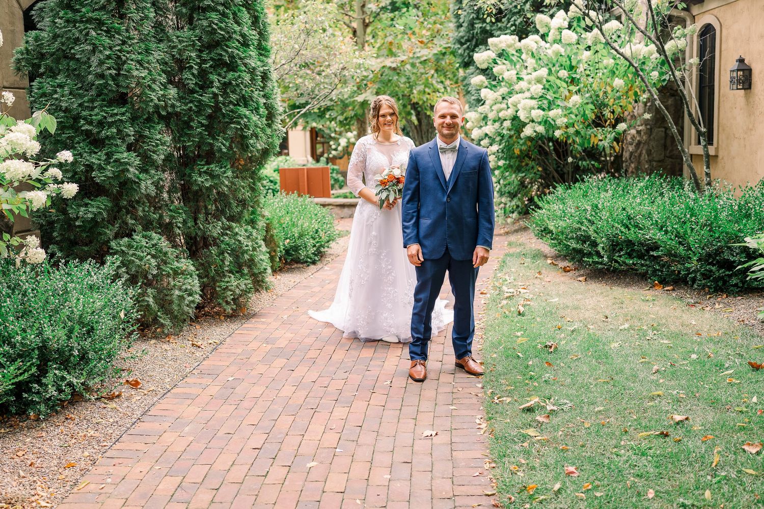 Bride and groom standing together on brick garden path surrounded by lush greenery.