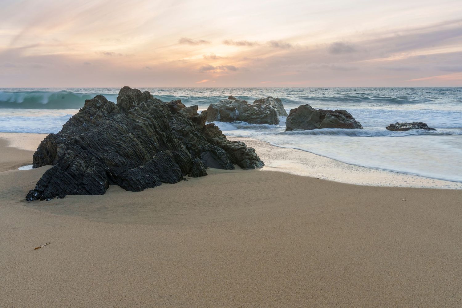 Rocky shoreline with dark boulders on sandy beach at sunset, gentle waves washing ashore under a pastel sky in Big Sur, California