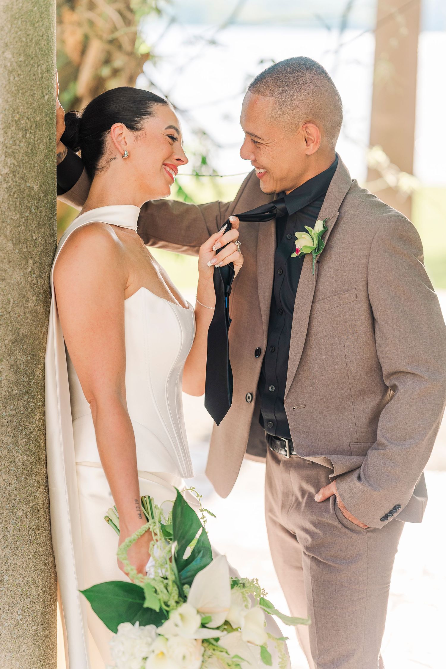 Bride in halter gown playfully adjusting groom's tie while holding white bouquet in outdoor setting.