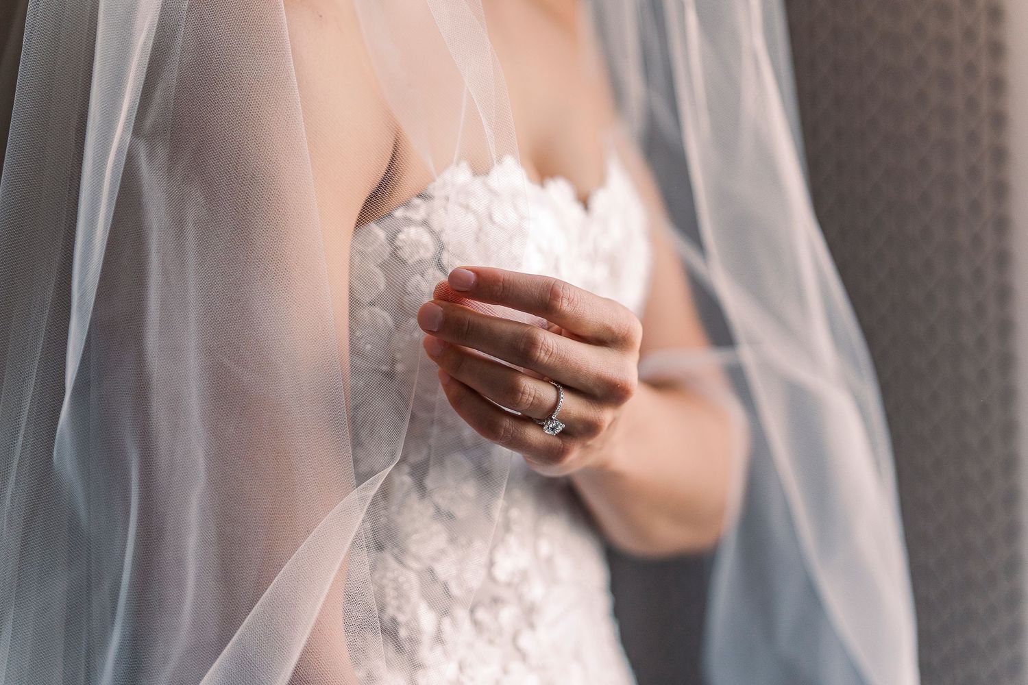 Bride in white lace wedding dress with veil, wearing a diamond ring, holding bouquet close to her chest.