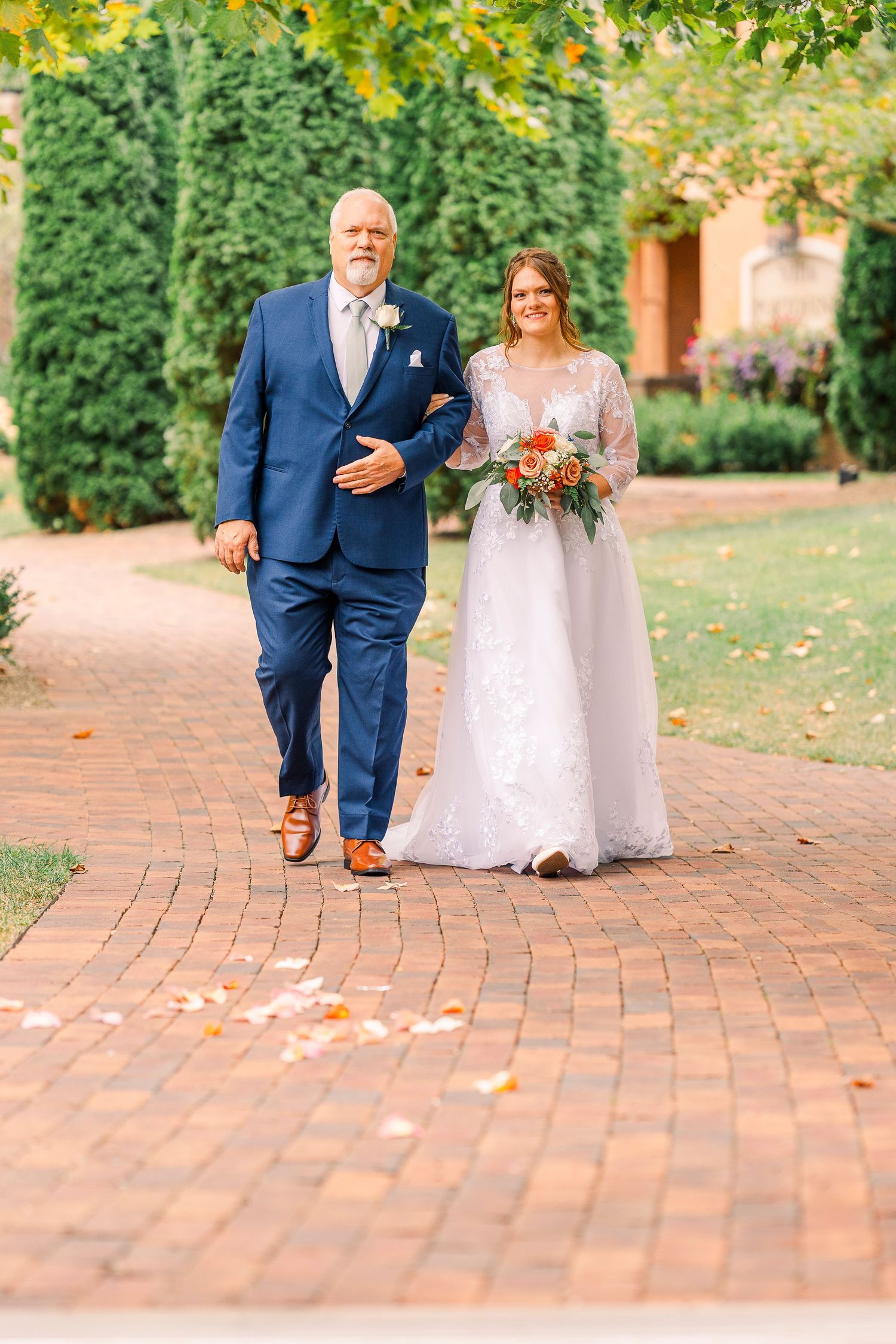 Bride in lace gown holding orange bouquet walks with older man in navy suit on brick path.