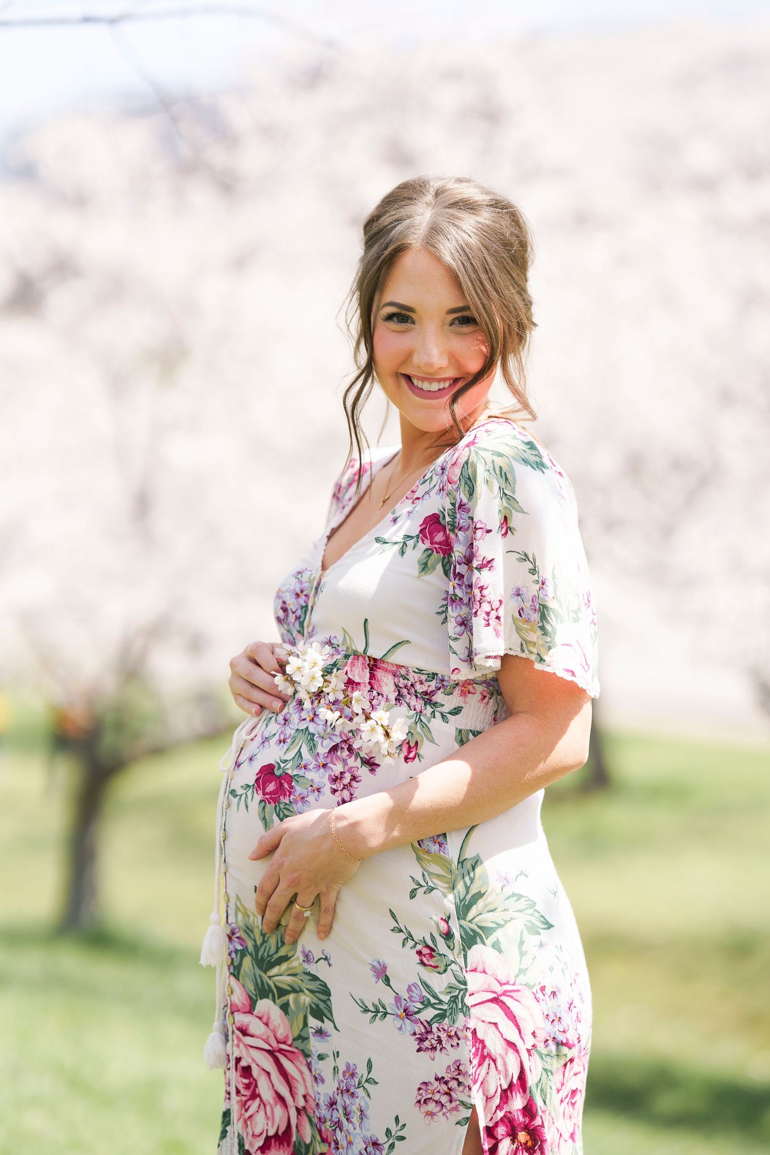 Smiling pregnant woman in floral dress cradles her baby bump outdoors among Cherry Blossoms in Cleveland, Ohio