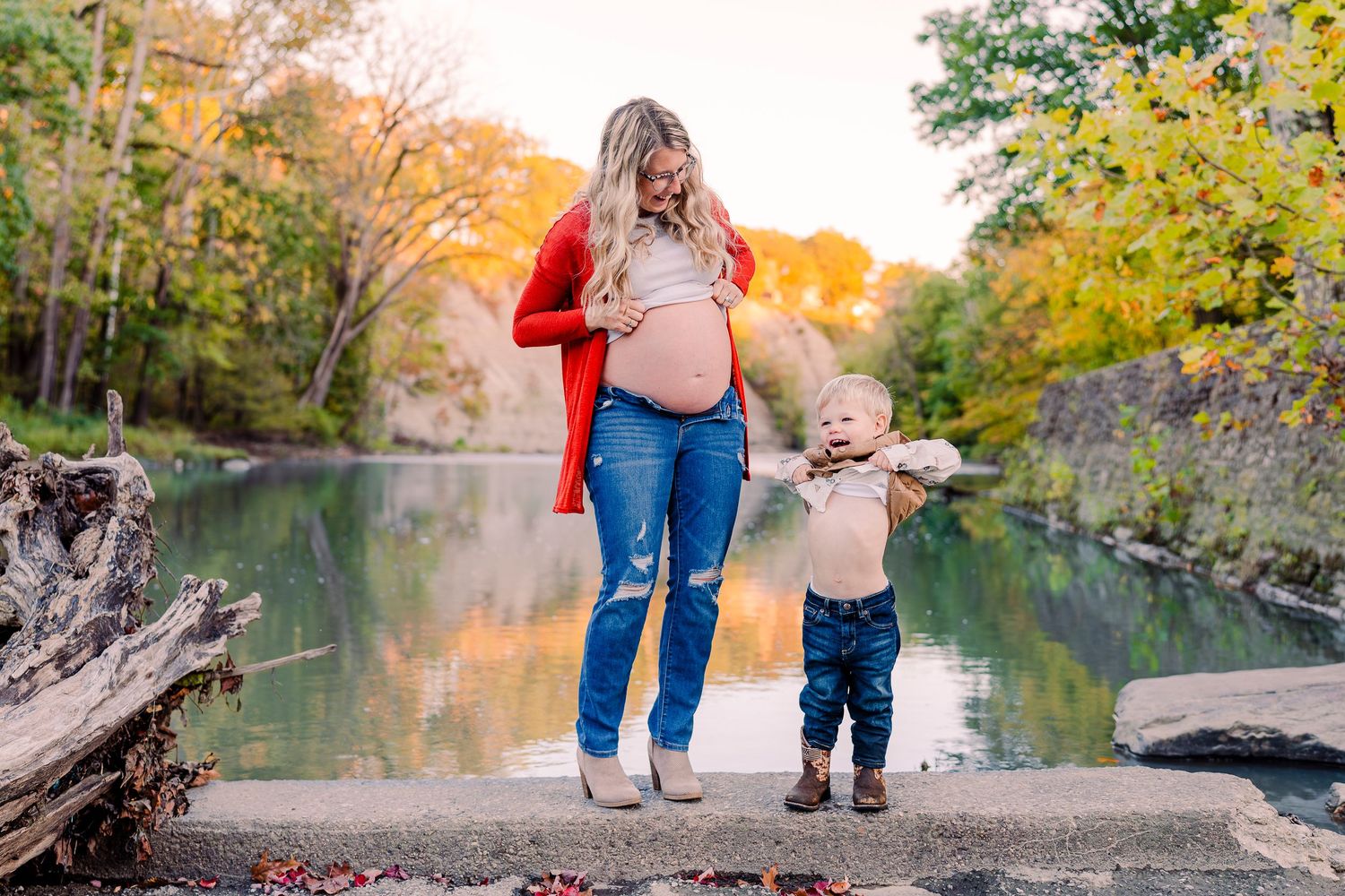 Pregnant mom in red cardigan poses with her young child by a beautiful fall foliage riverside setting in Rocky River, Ohio