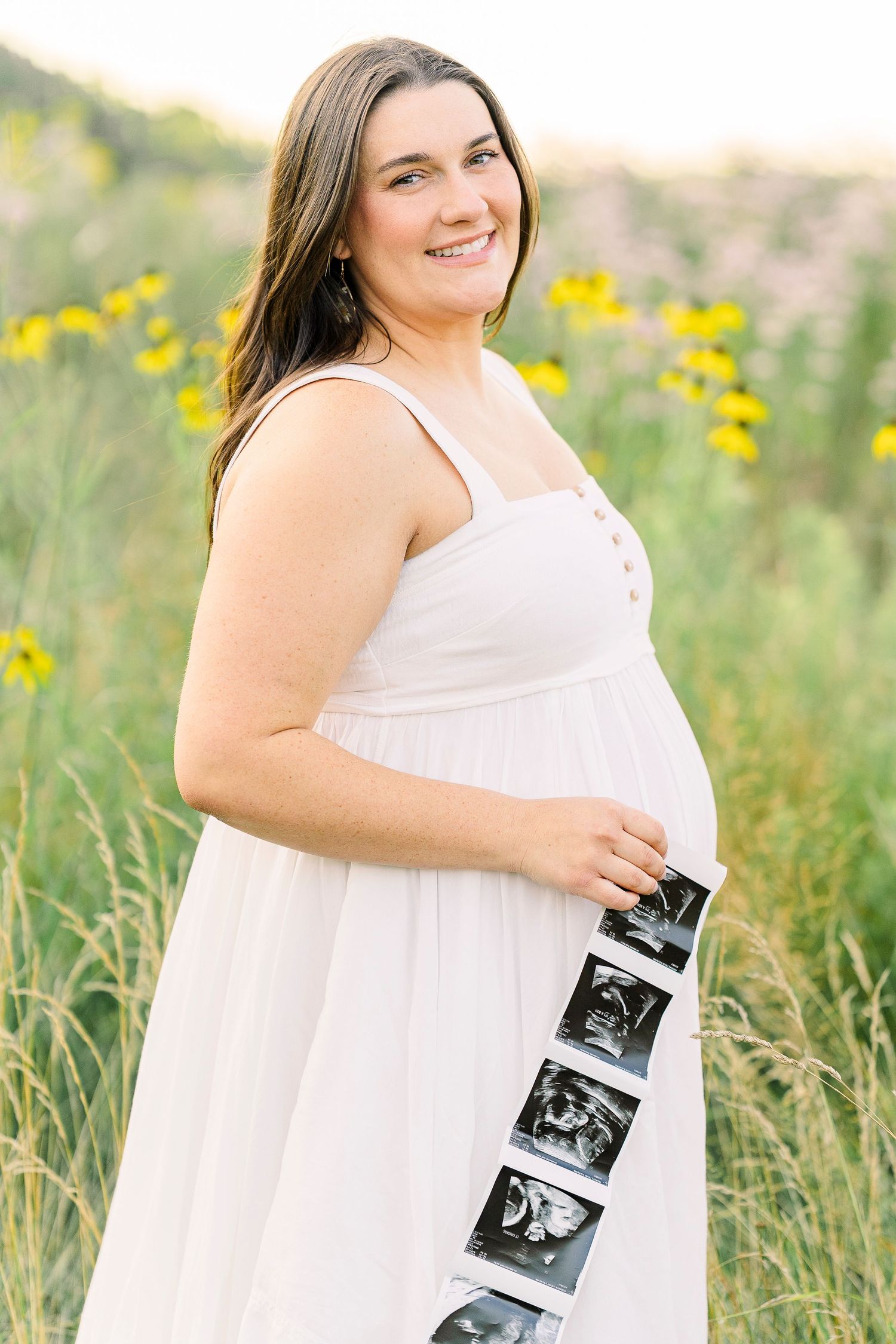 A smiling pregnant woman in a white dress holds ultrasound photos in a field of yellow wildflowers.