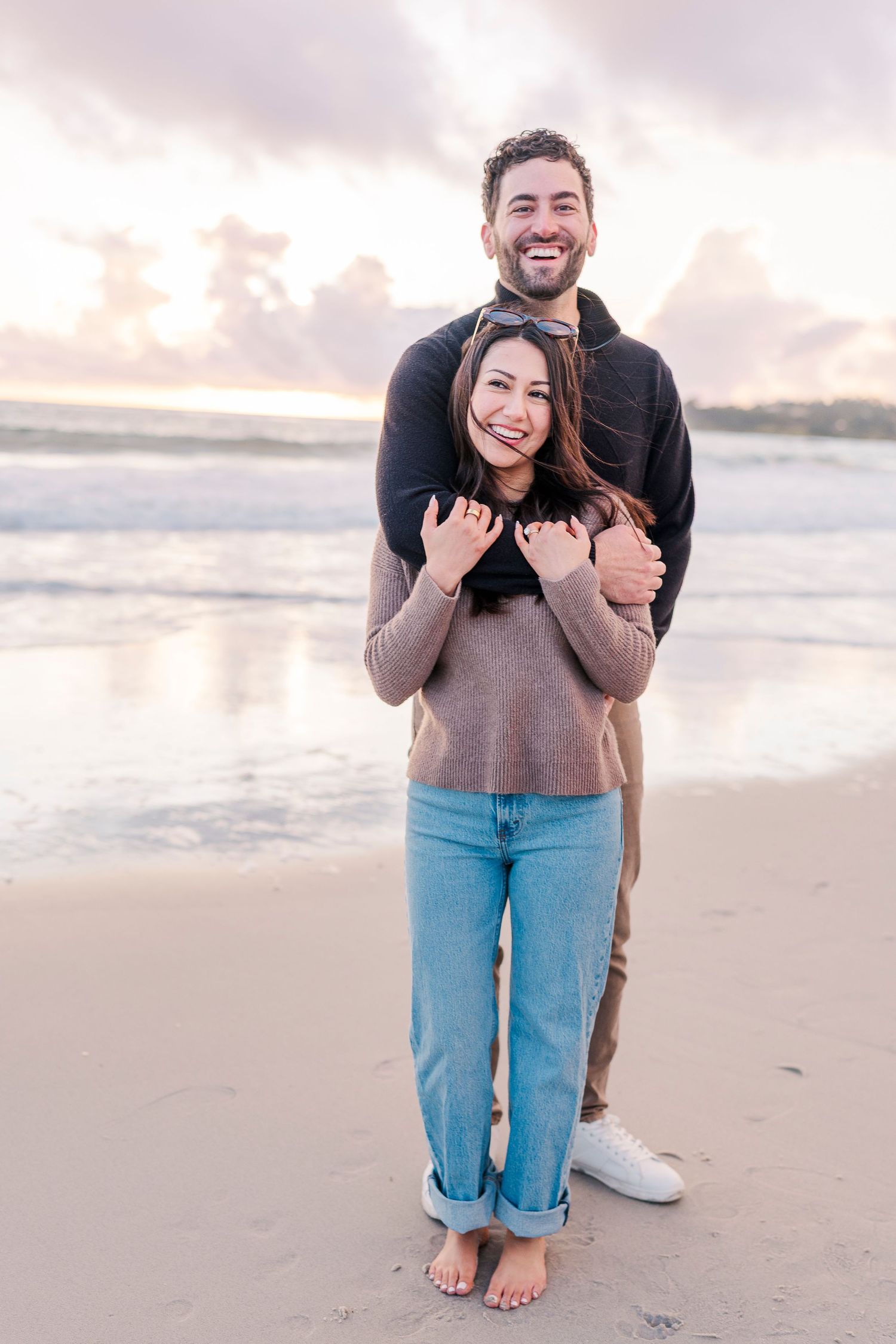 A couple embraces on a beach at sunset, the man hugging the woman from behind as waves reflect pastel skies on Carmel beach in California