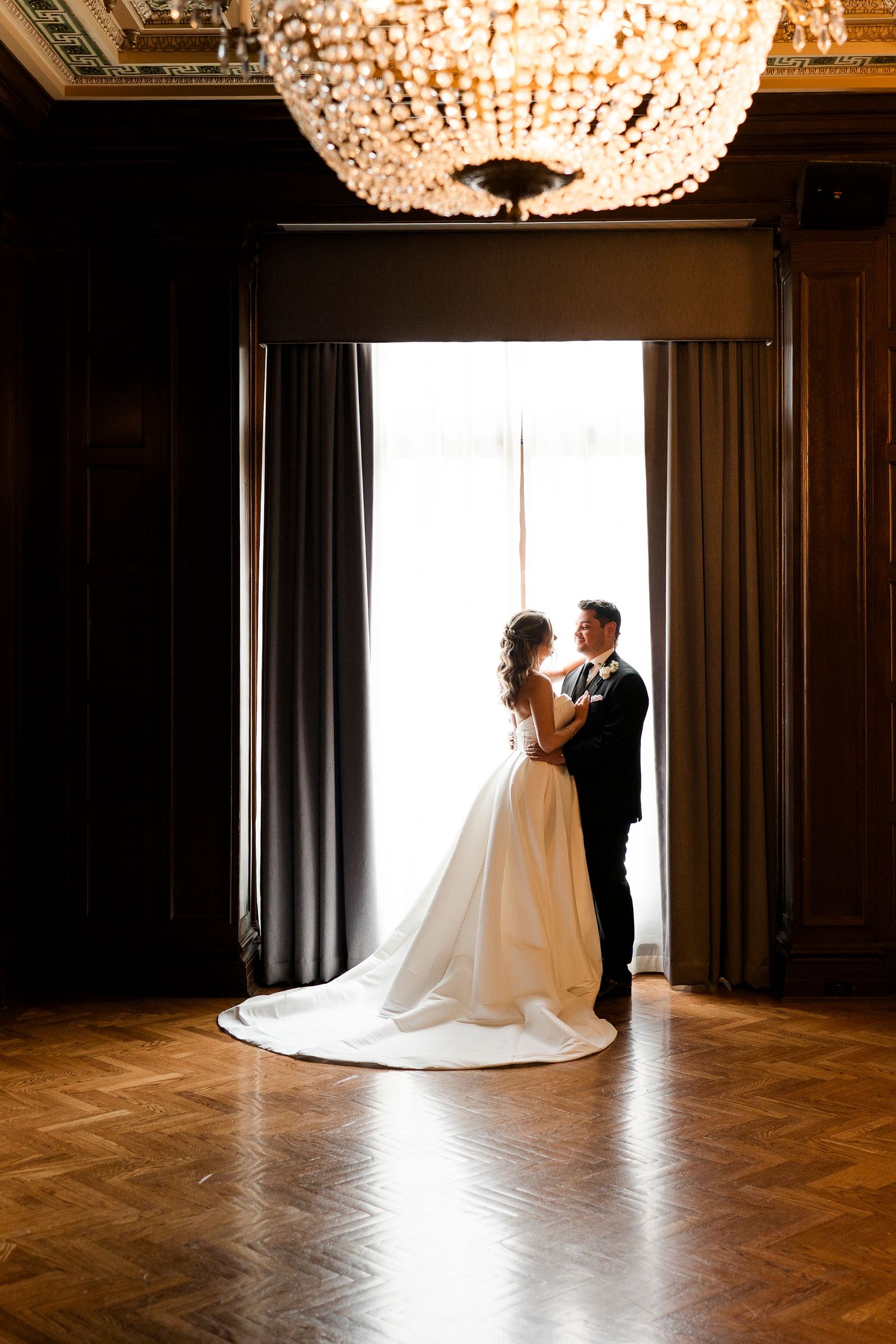 Bride and groom silhouetted in elegant ballroom doorway with crystal chandelier and herringbone wood floor at the Columbus Athletic club