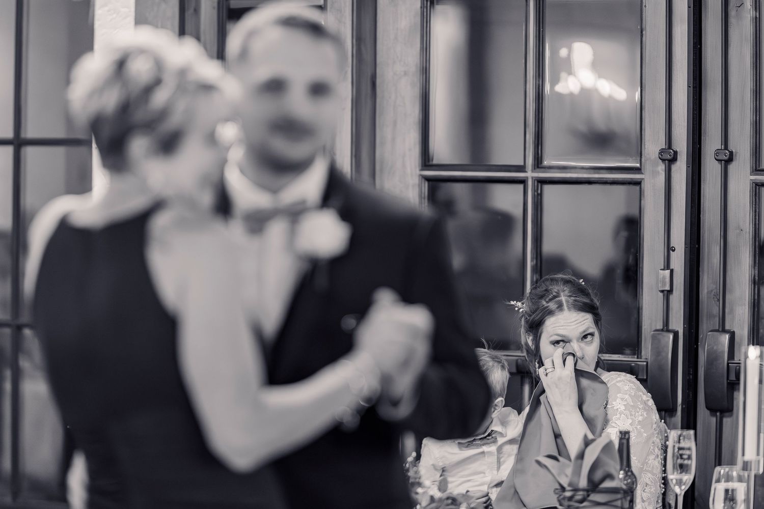 Bride wipes tears watching groom dance with his mother at elegant wedding reception in black and white.