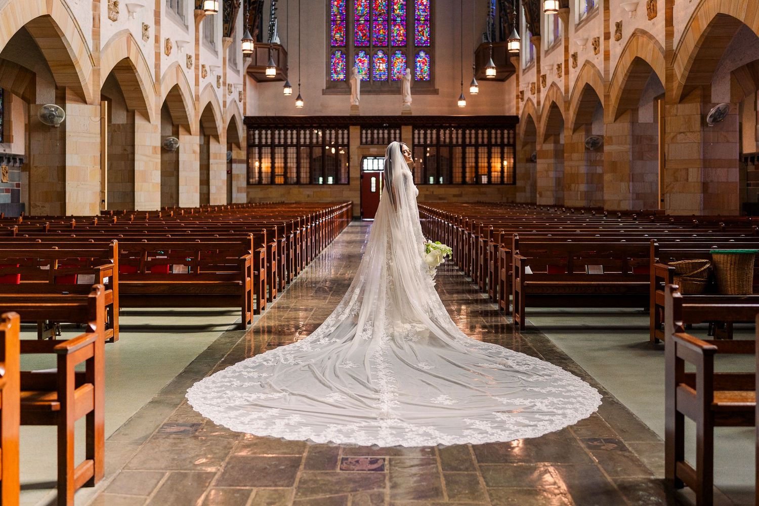 Bride in white gown with long cathedral veil walks down church aisle toward altar with stained glass windows.