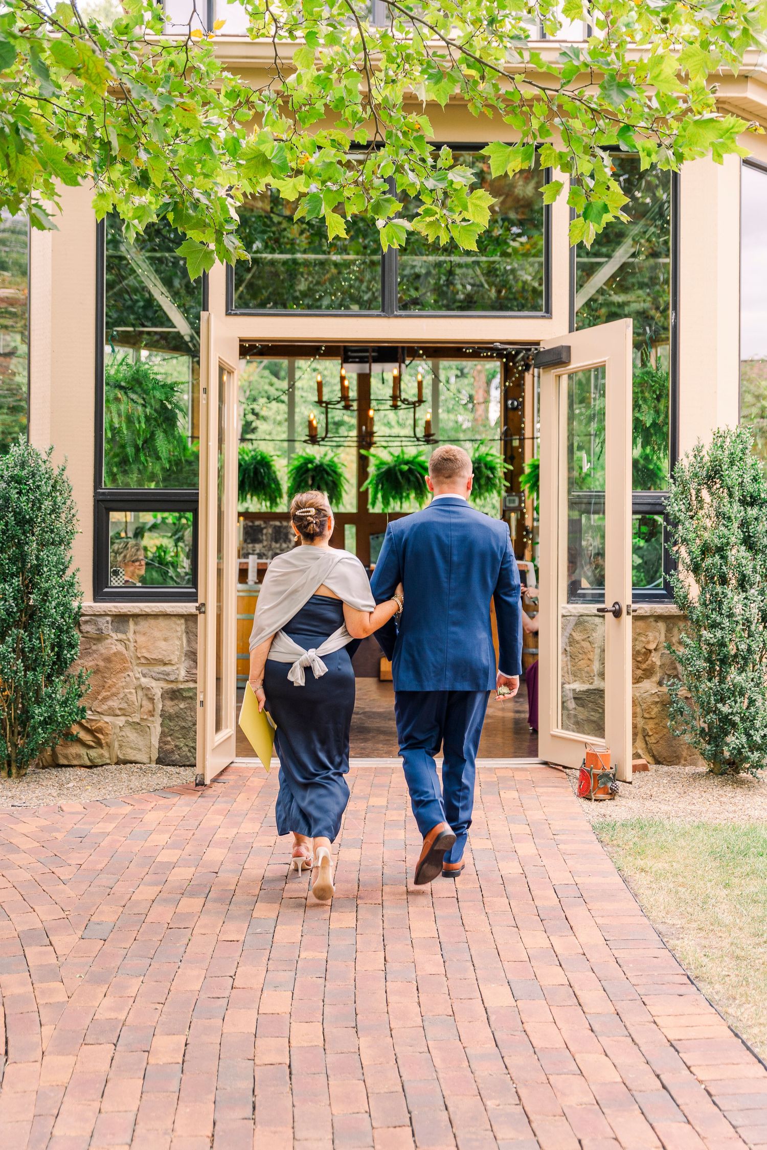 Couple walks arm-in-arm toward glass greenhouse venue entrance framed by lush green foliage.