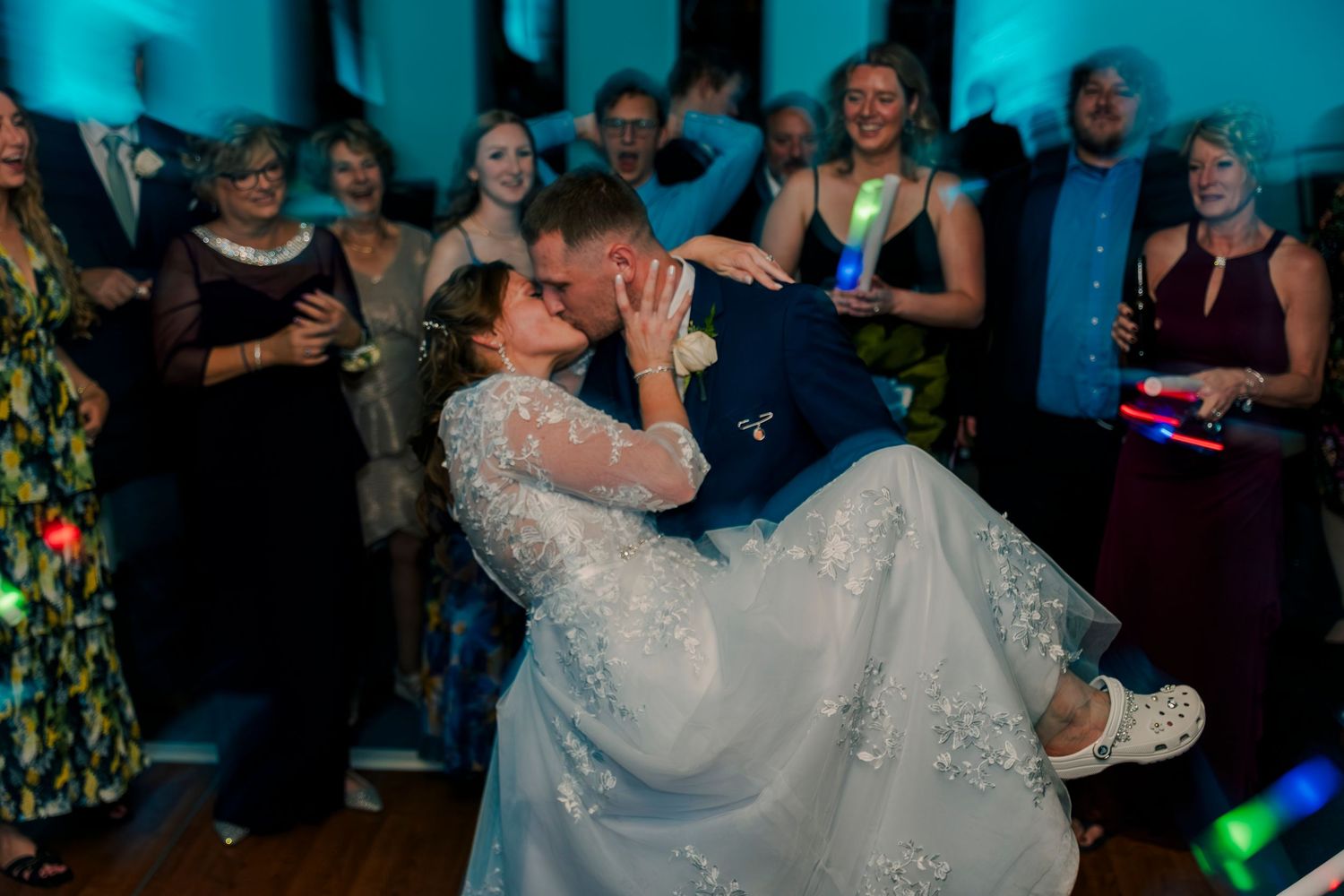 Groom dips bride for a romantic kiss on the dance floor surrounded by cheering wedding guests.